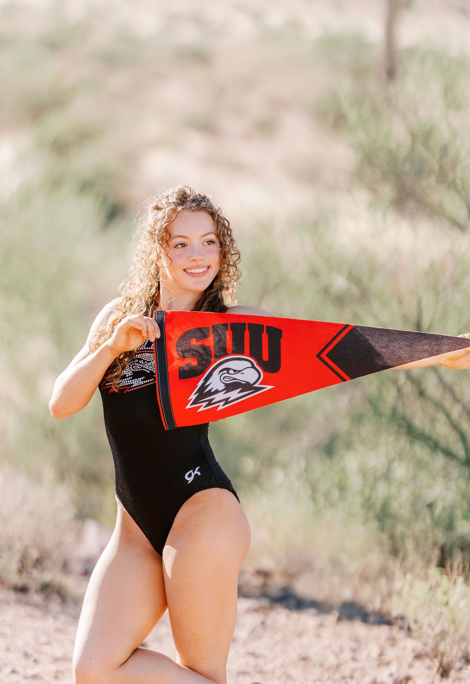 A gymnast in a black leotard poses outdoors while holding a red SUU athletic pennant.