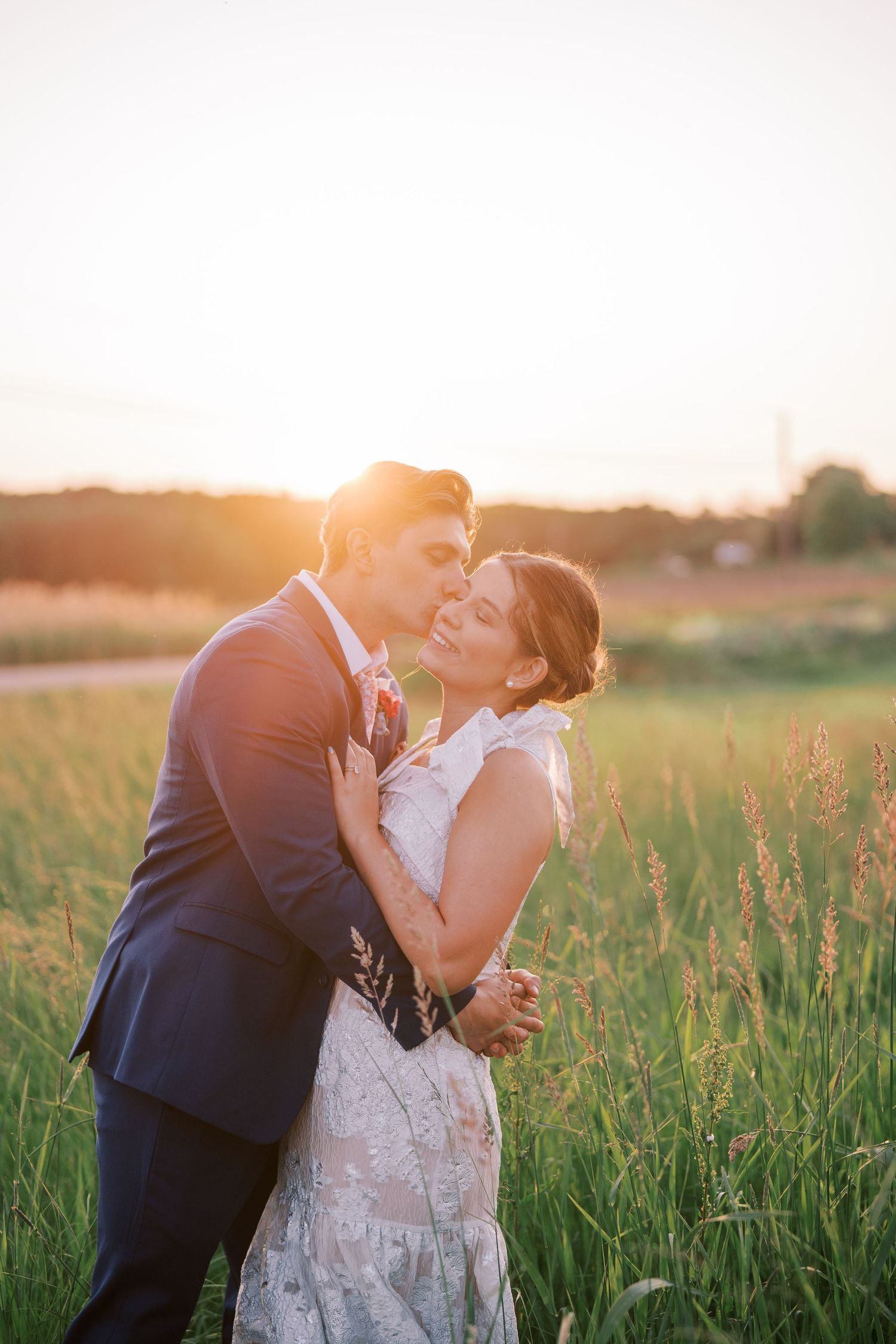 A wedding couple embraces romantically in a golden-lit meadow during sunset.
