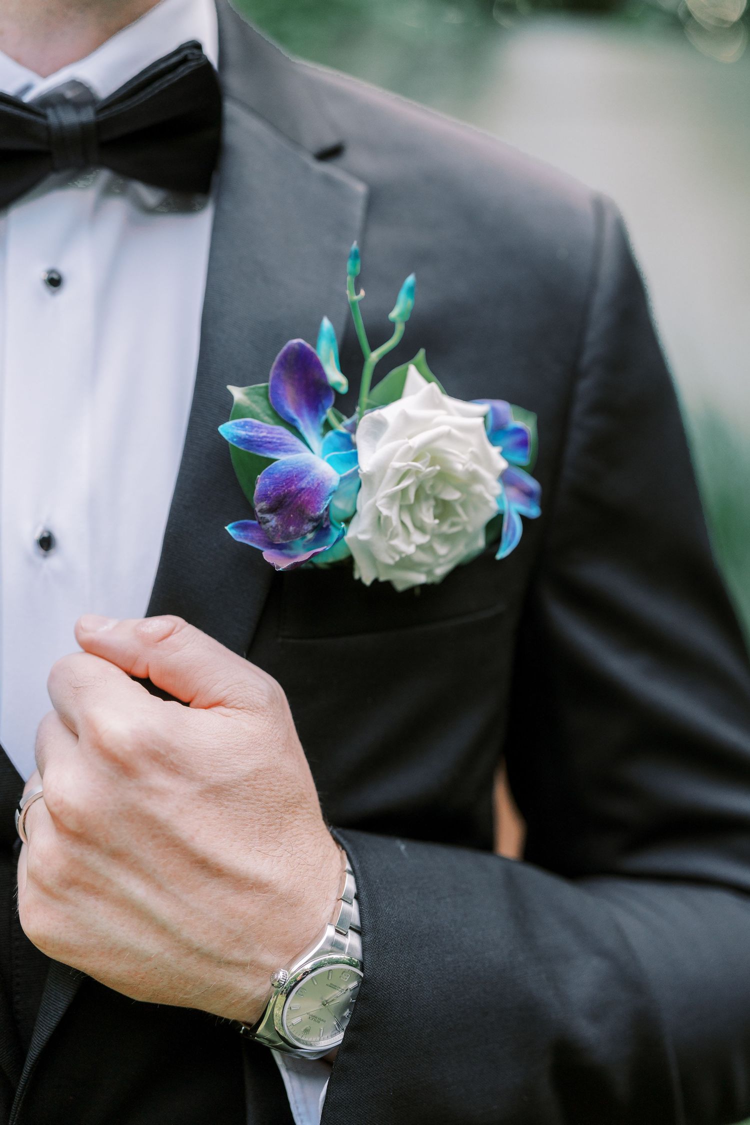 A close-up of a black tuxedo lapel with blue orchid and white rose boutonniere and dress watch visible.