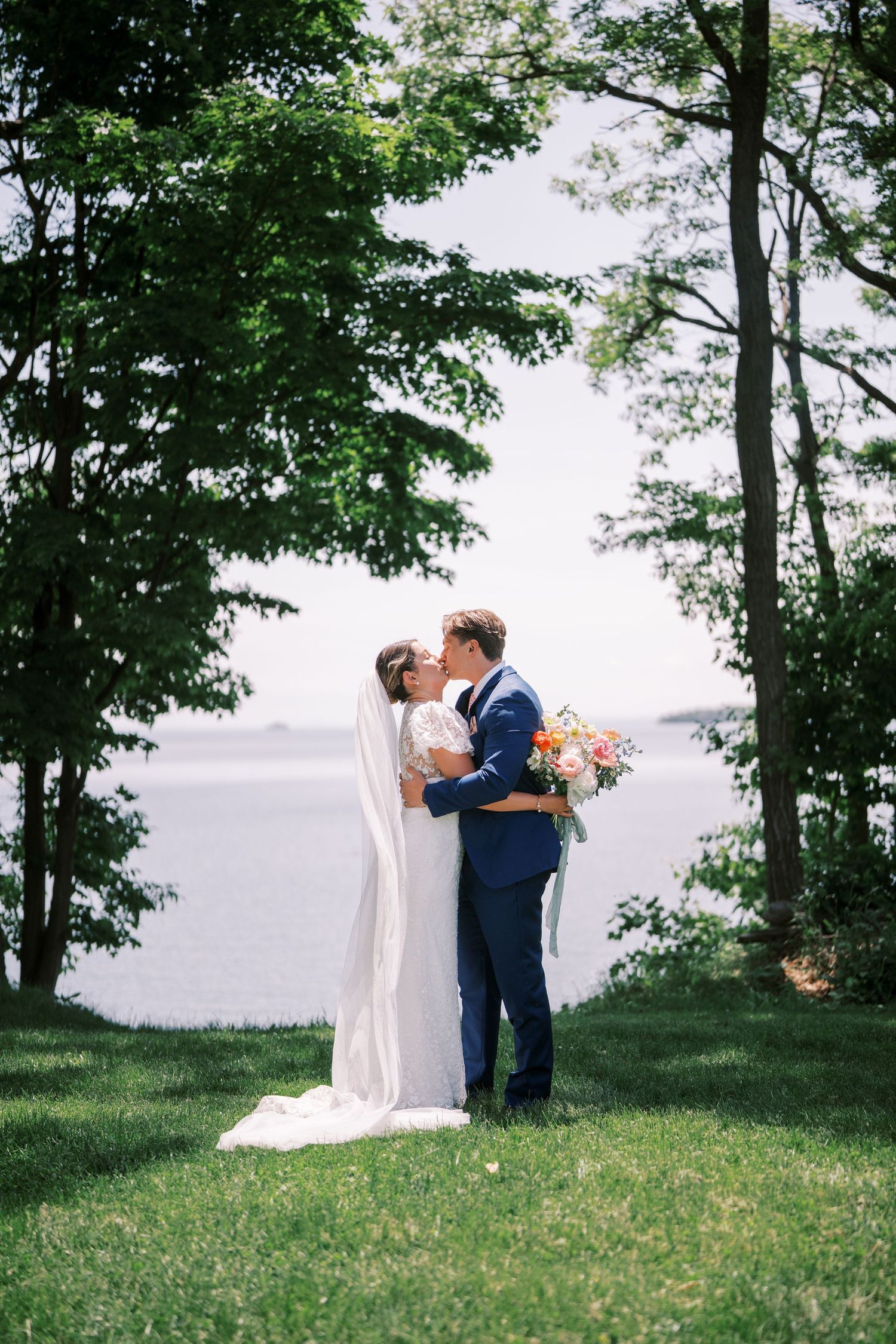 A couple shares an intimate wedding moment by a lake, surrounded by lush trees on a manicured lawn.
