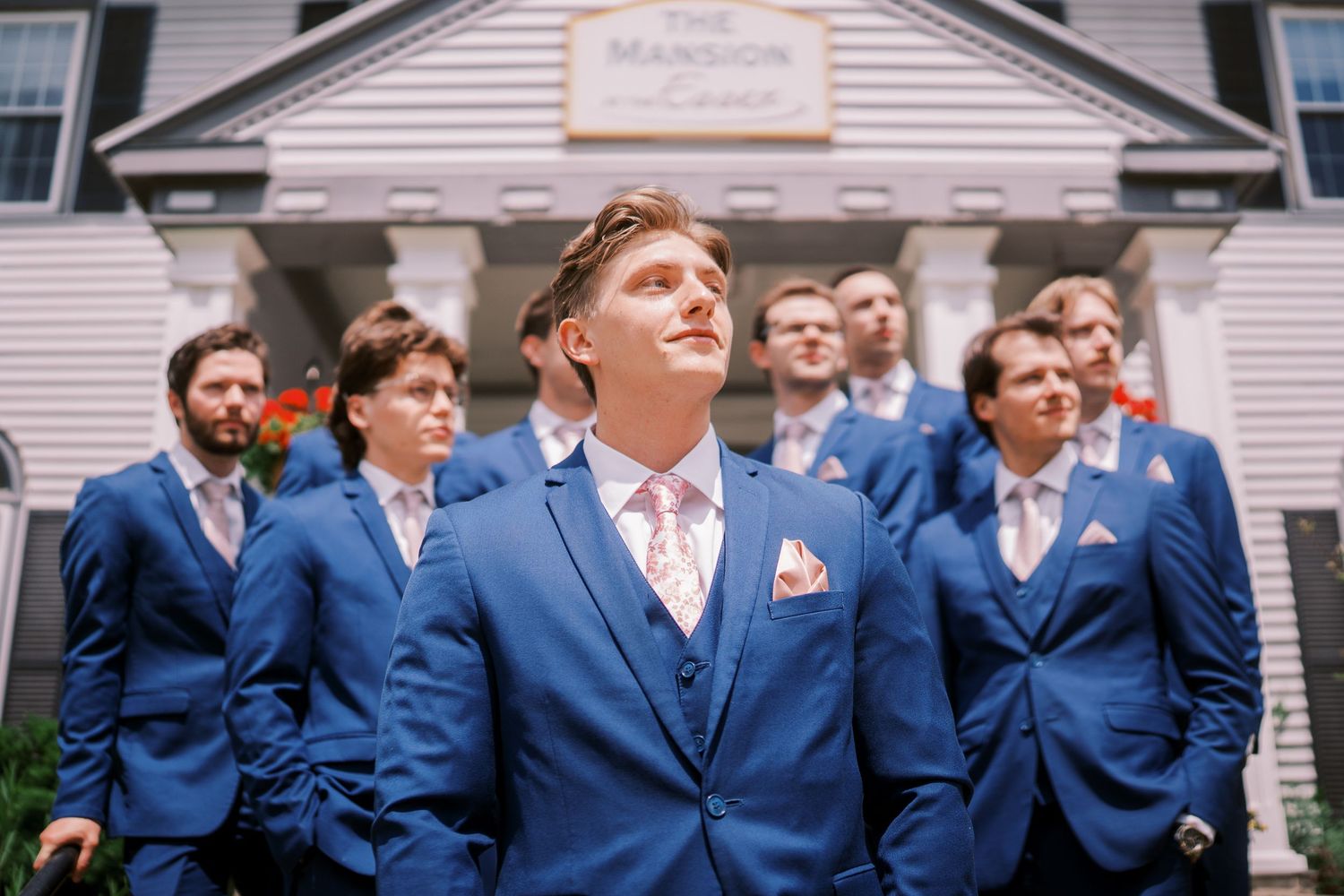 Groomsmen in matching navy blue suits stand in line outside a white building for a wedding photo.