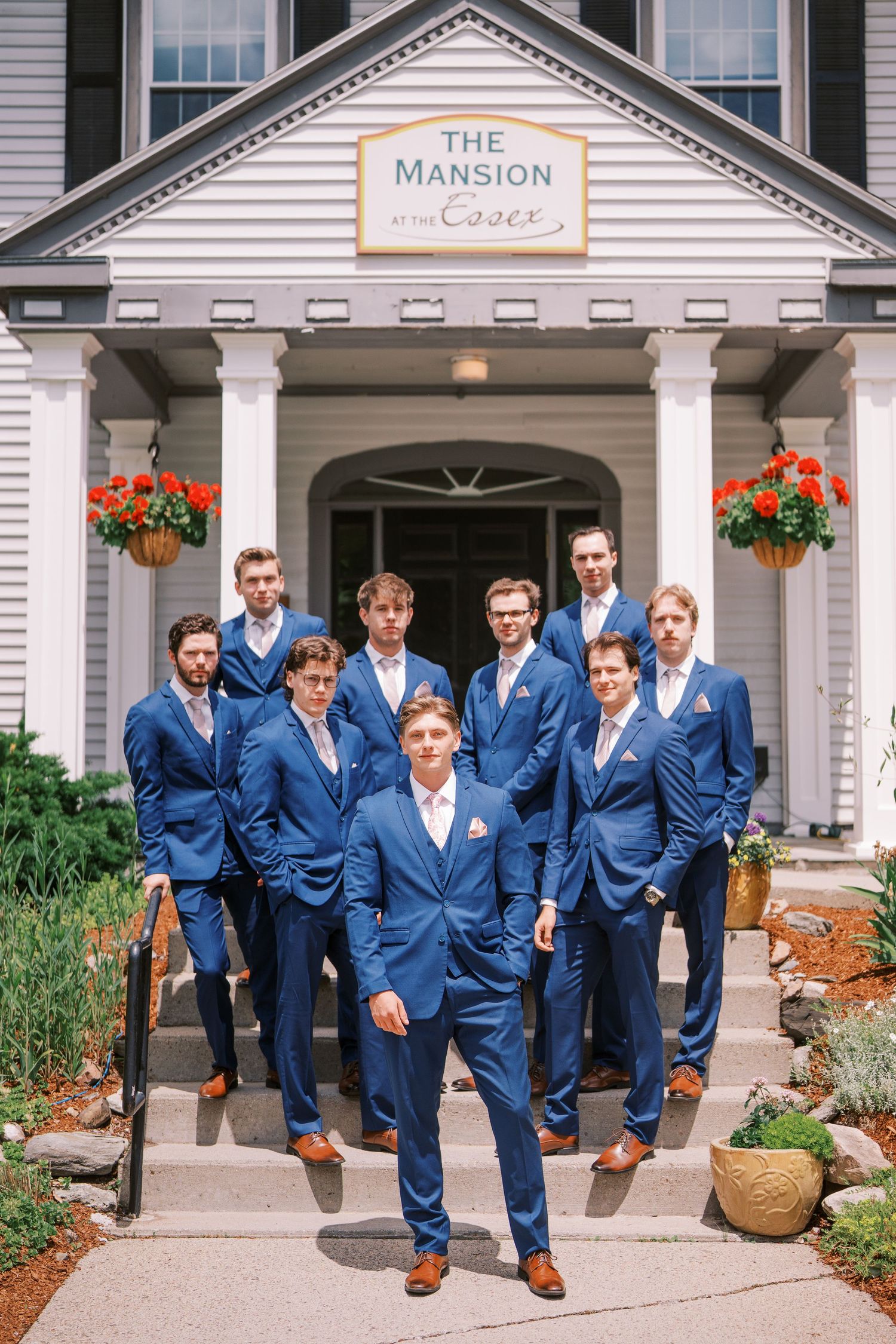 A group of groomsmen in matching navy blue suits pose on the steps of The Mansion wedding venue with bright floral decorations.