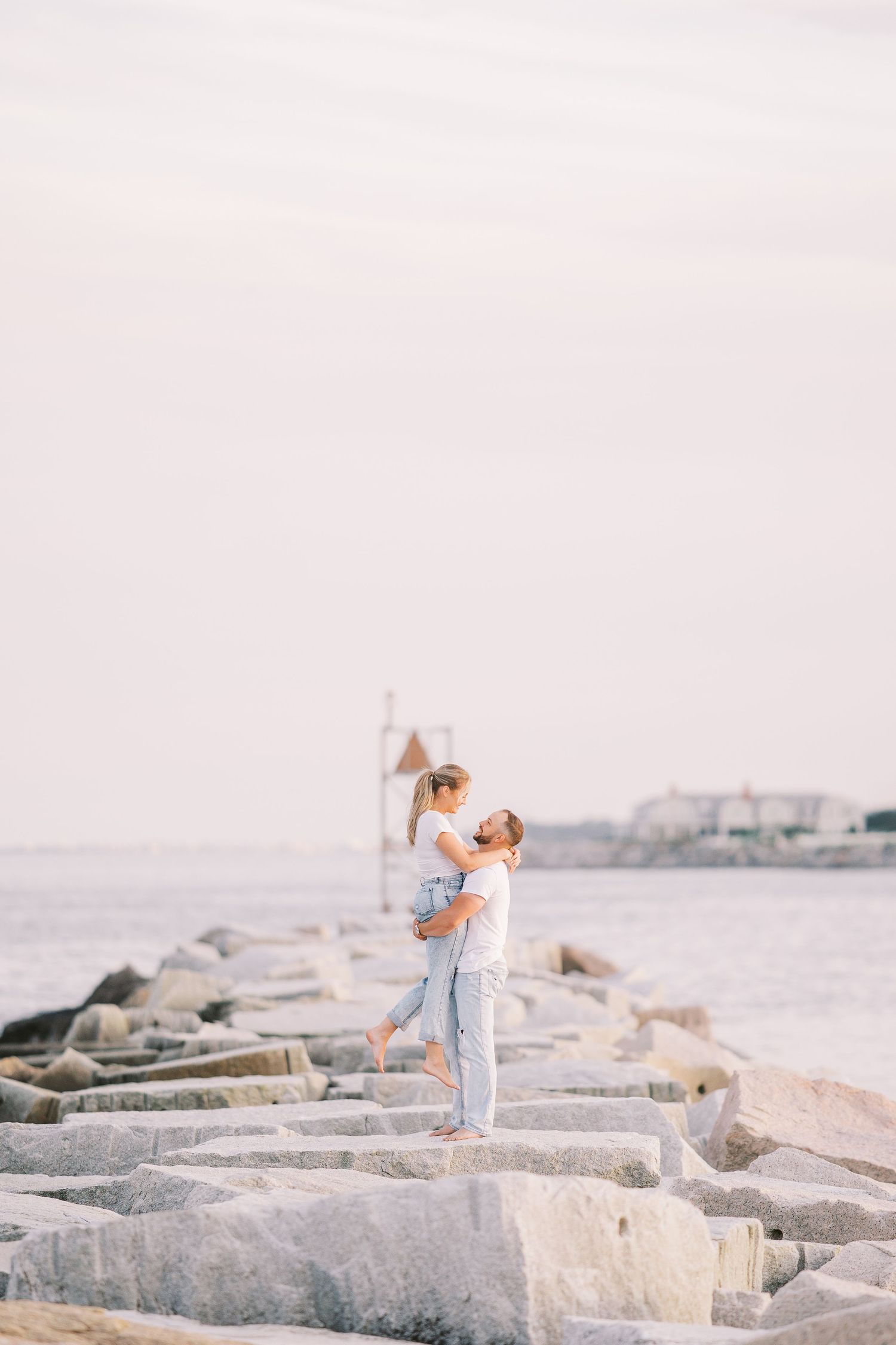 Person in light blue outfit walks along coastal rock jetty while holding a camera on cloudy day by the ocean.