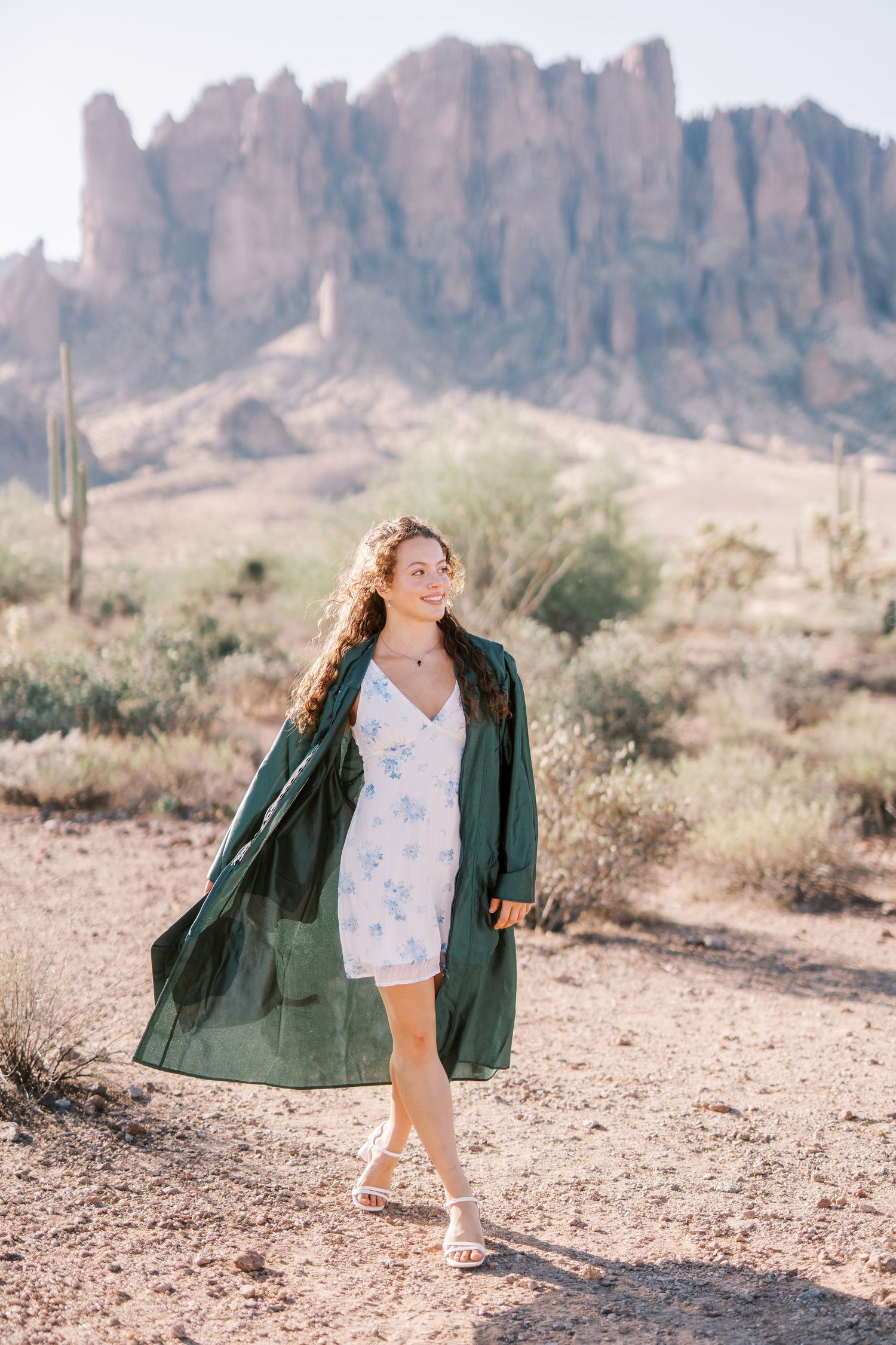 A person in a white dress and green coat walks through a desert landscape with mountains and saguaro cactus in the background.