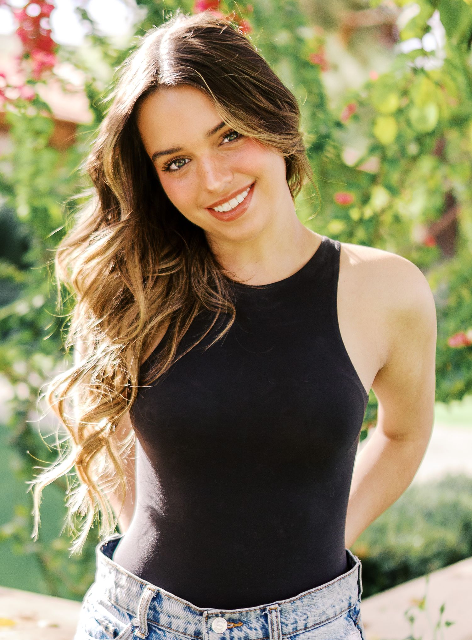 A lifestyle portrait in a garden setting with wavy brown hair, black sleeveless top and denim jeans.