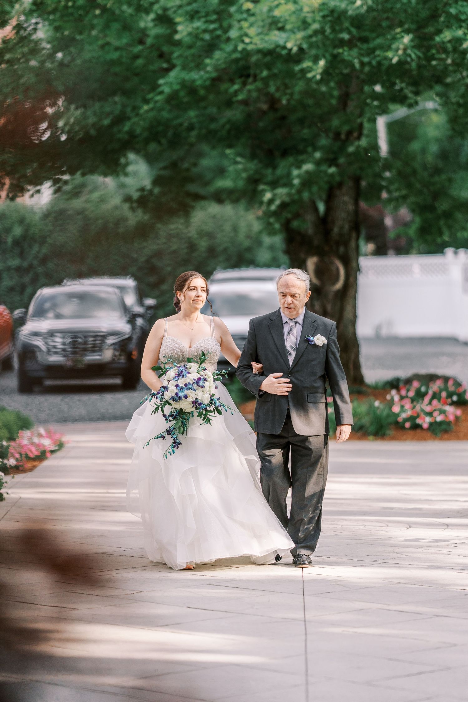 A bride walking down an outdoor pathway with someone escorting her on a wedding day.