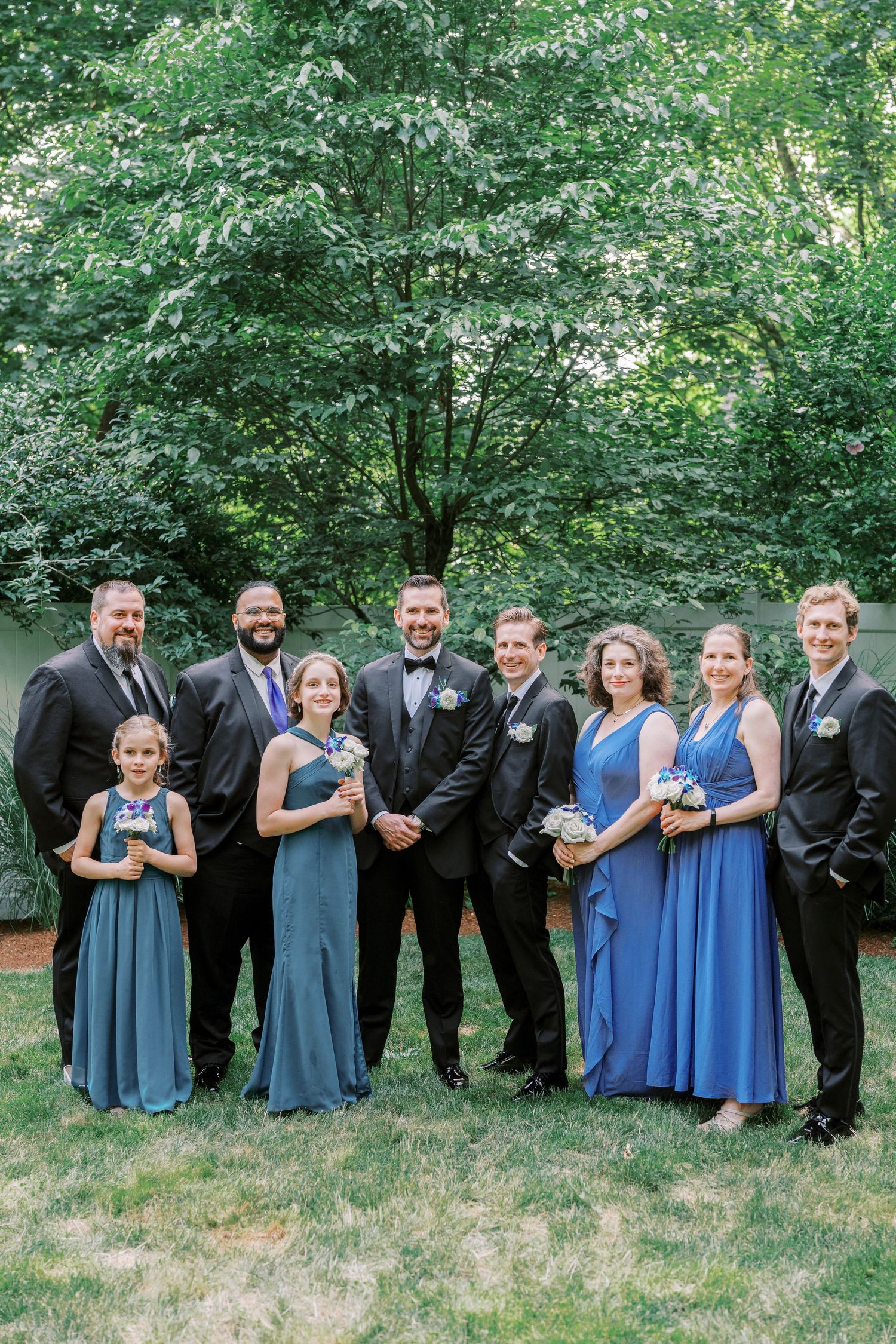 Wedding party in coordinated navy and blue formal attire poses outdoors against lush green foliage.
