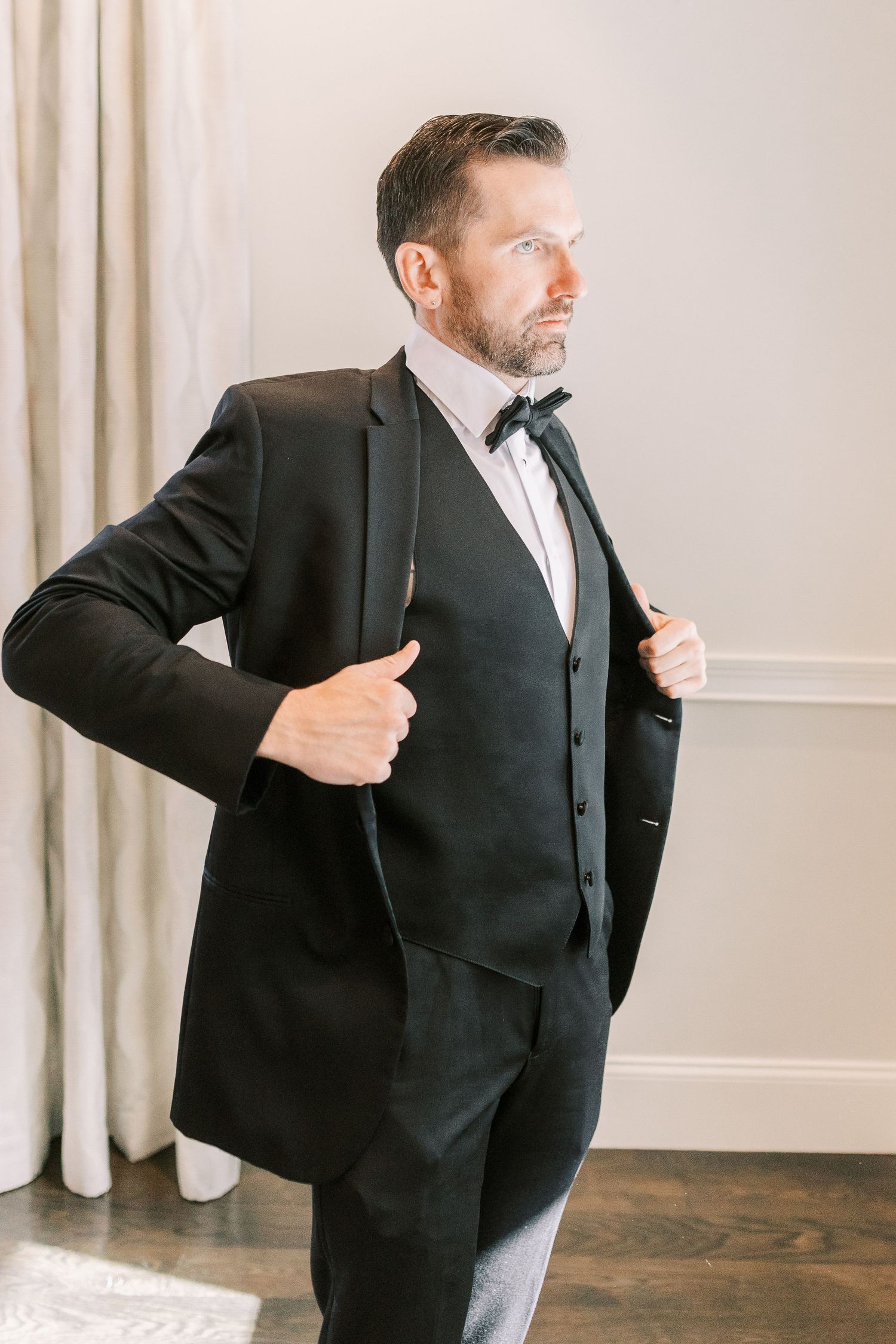 A well-dressed person in a classic black three-piece tuxedo with bow tie adjusts their jacket while standing indoors.