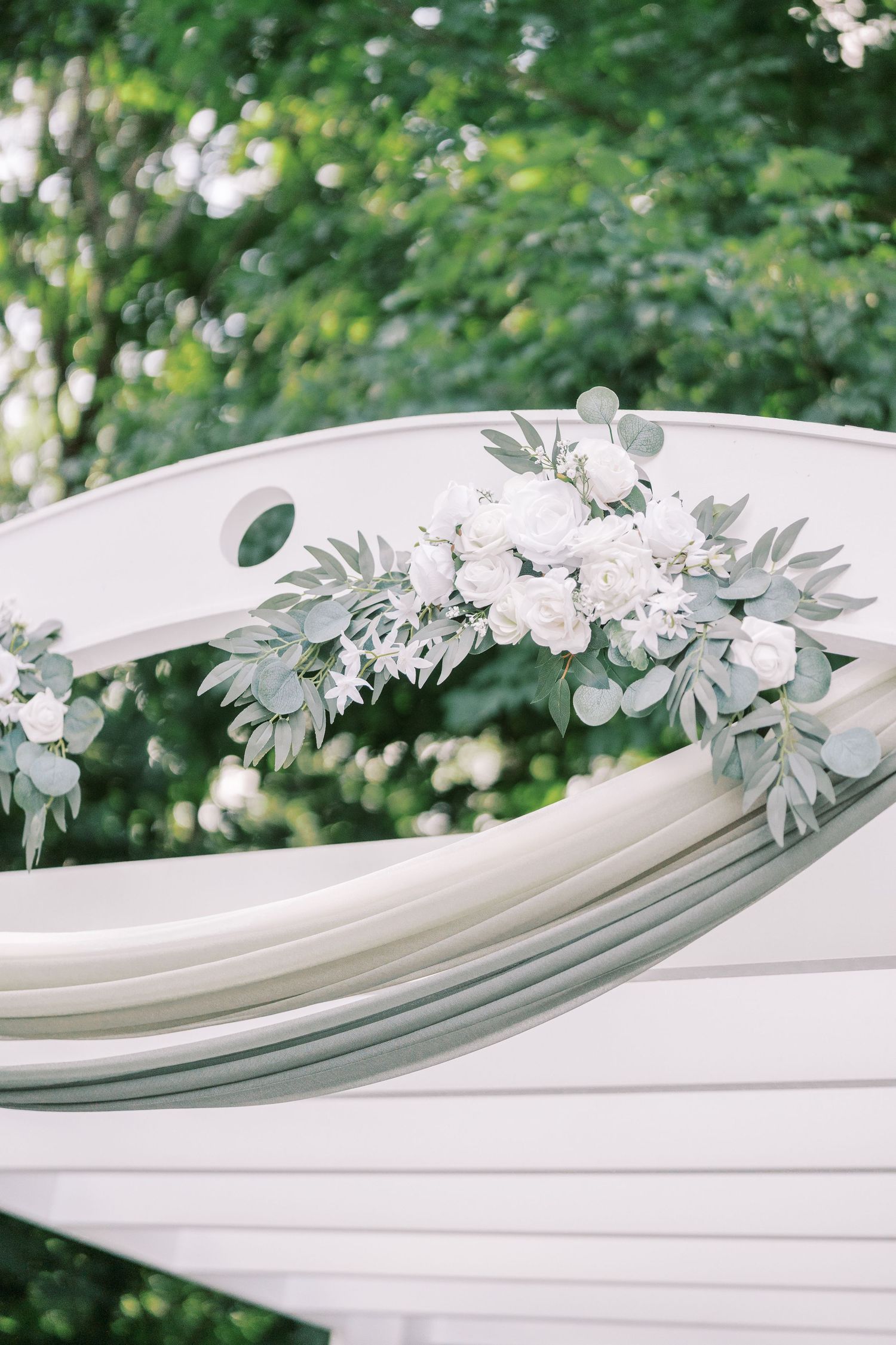 White wedding arch decorated with soft white roses and eucalyptus greenery against a blurred outdoor garden background.