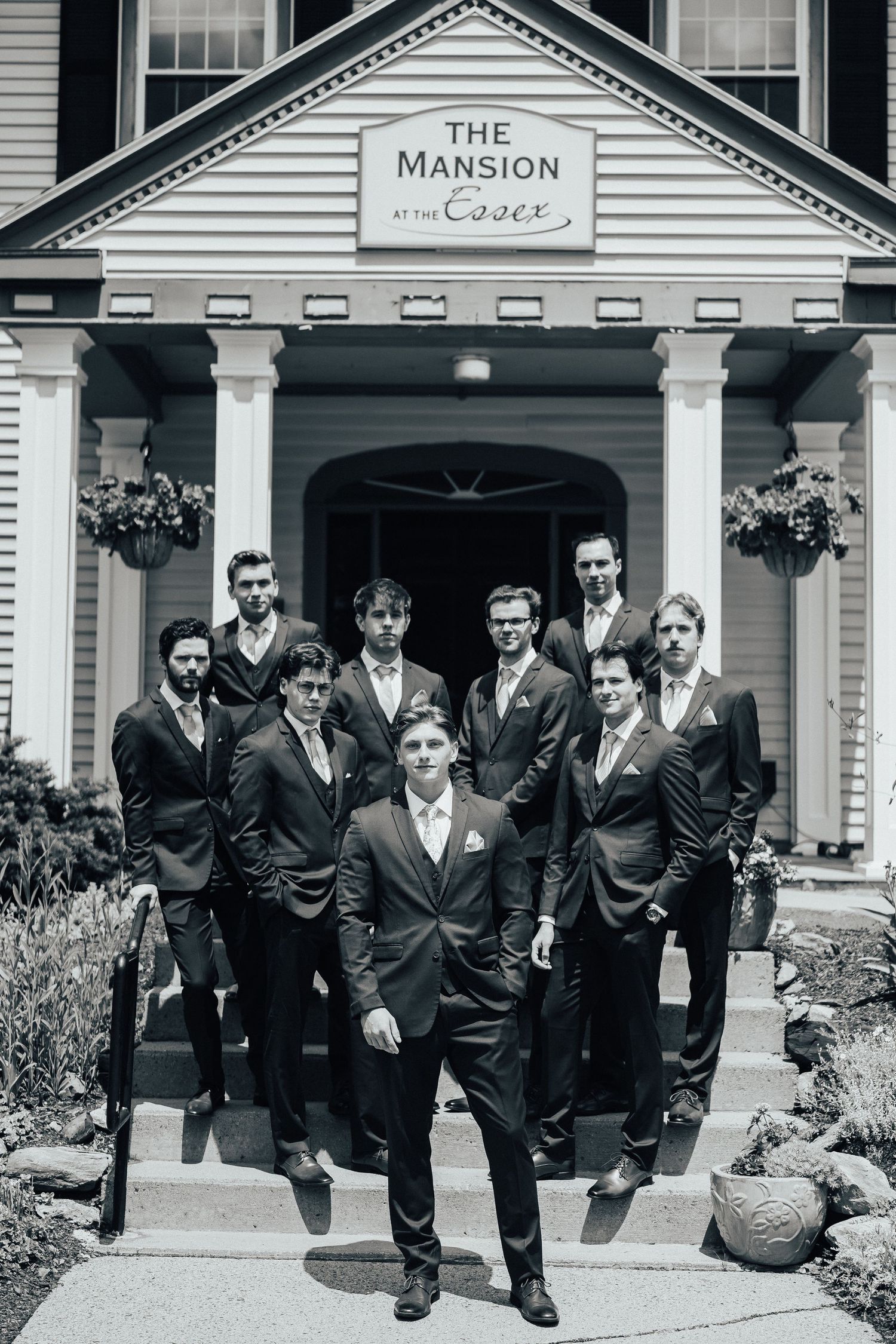 A group of groomsmen in dark suits pose together on the steps of The Mansion wedding venue in black and white.