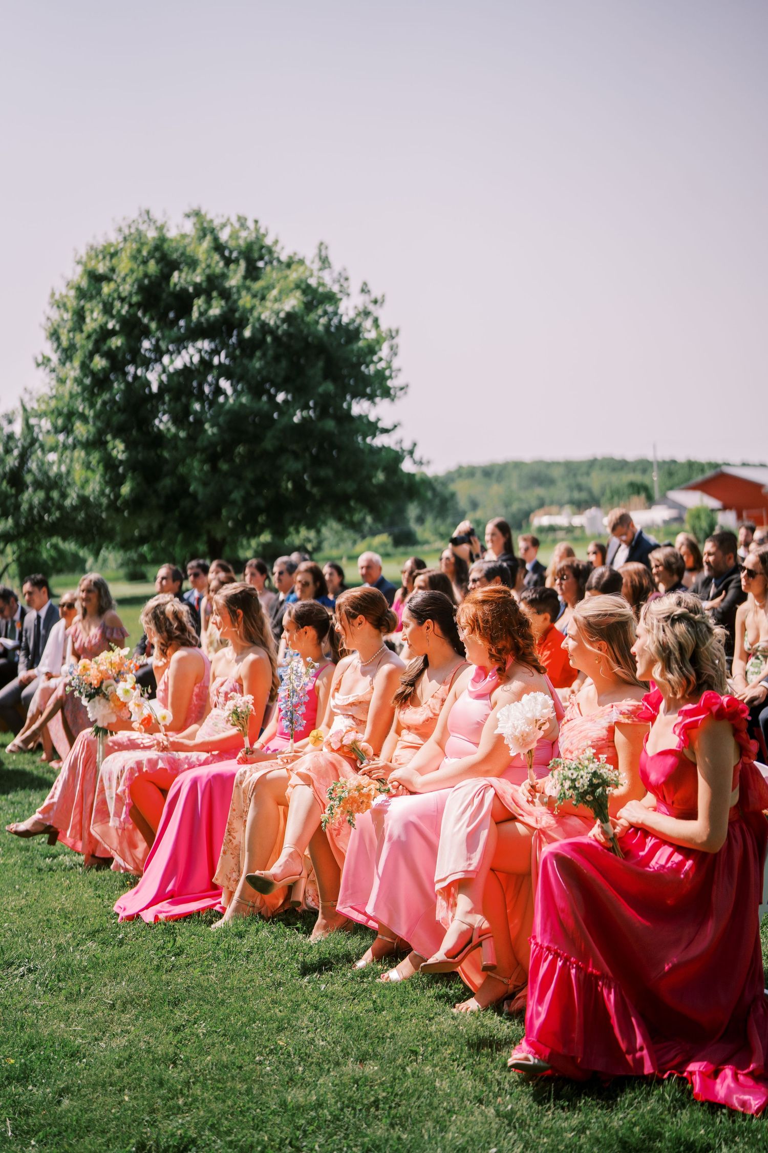 A row of bridesmaids wearing pink and coral dresses sit on chairs outdoors during a wedding ceremony.