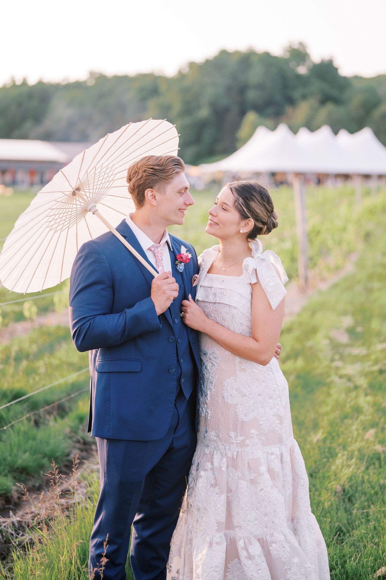 A happy couple in wedding attire stands in a field holding a white parasol during a sunny outdoor celebration.