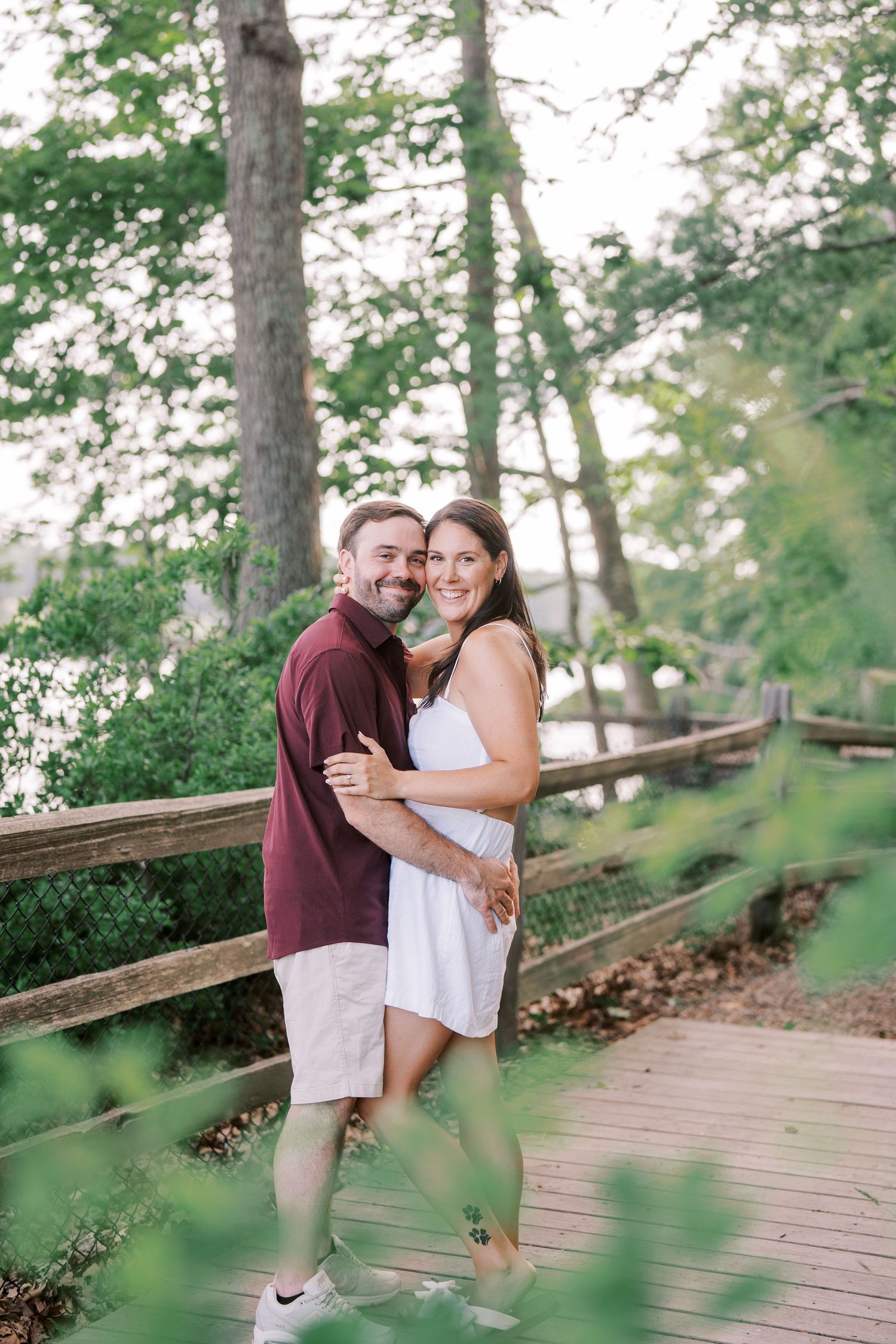 A romantic couple embraces on a wooden boardwalk surrounded by lush green trees and natural scenery.