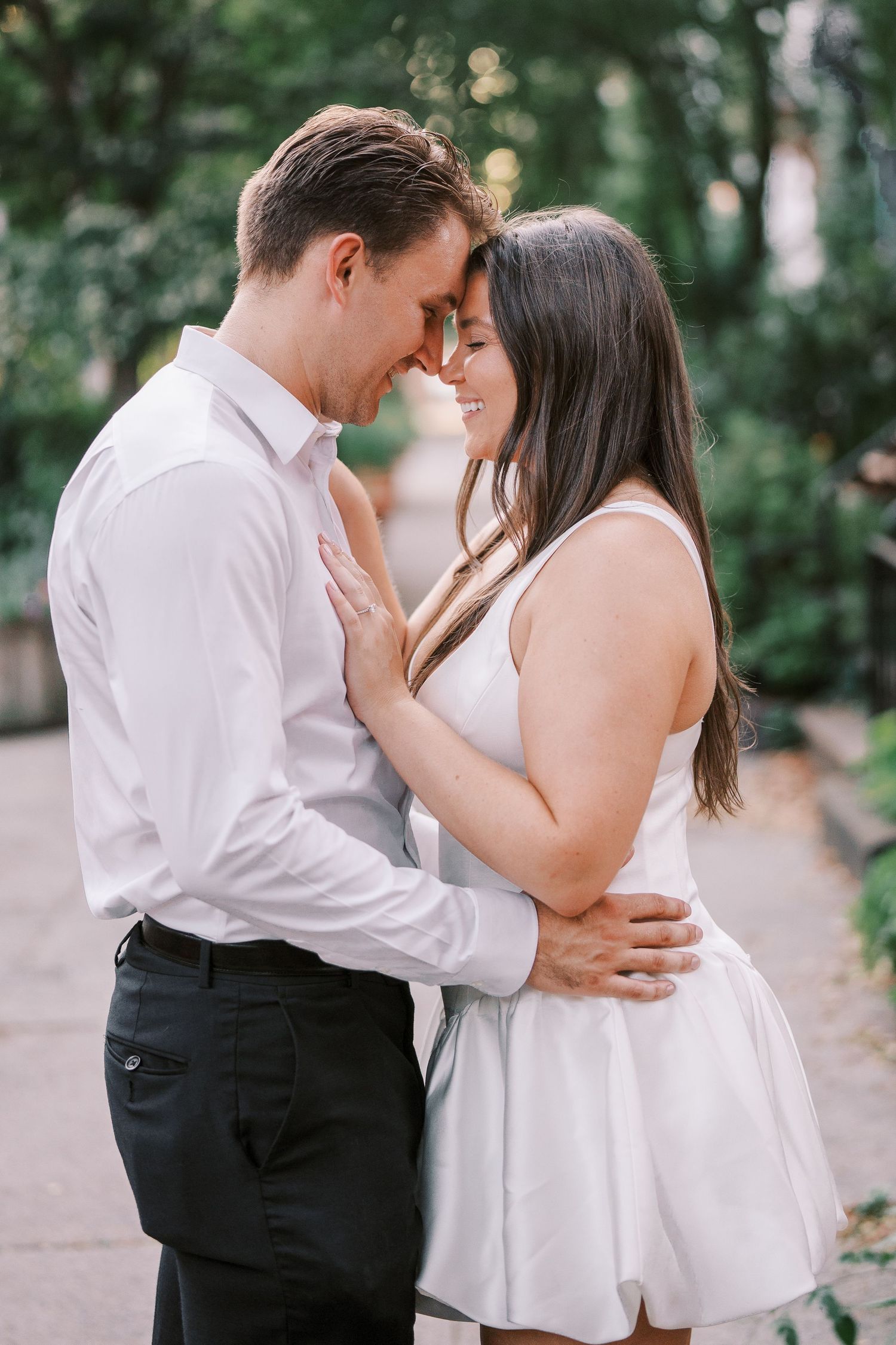A romantic couple in white clothing share an intimate moment outdoors among trees with soft lighting.