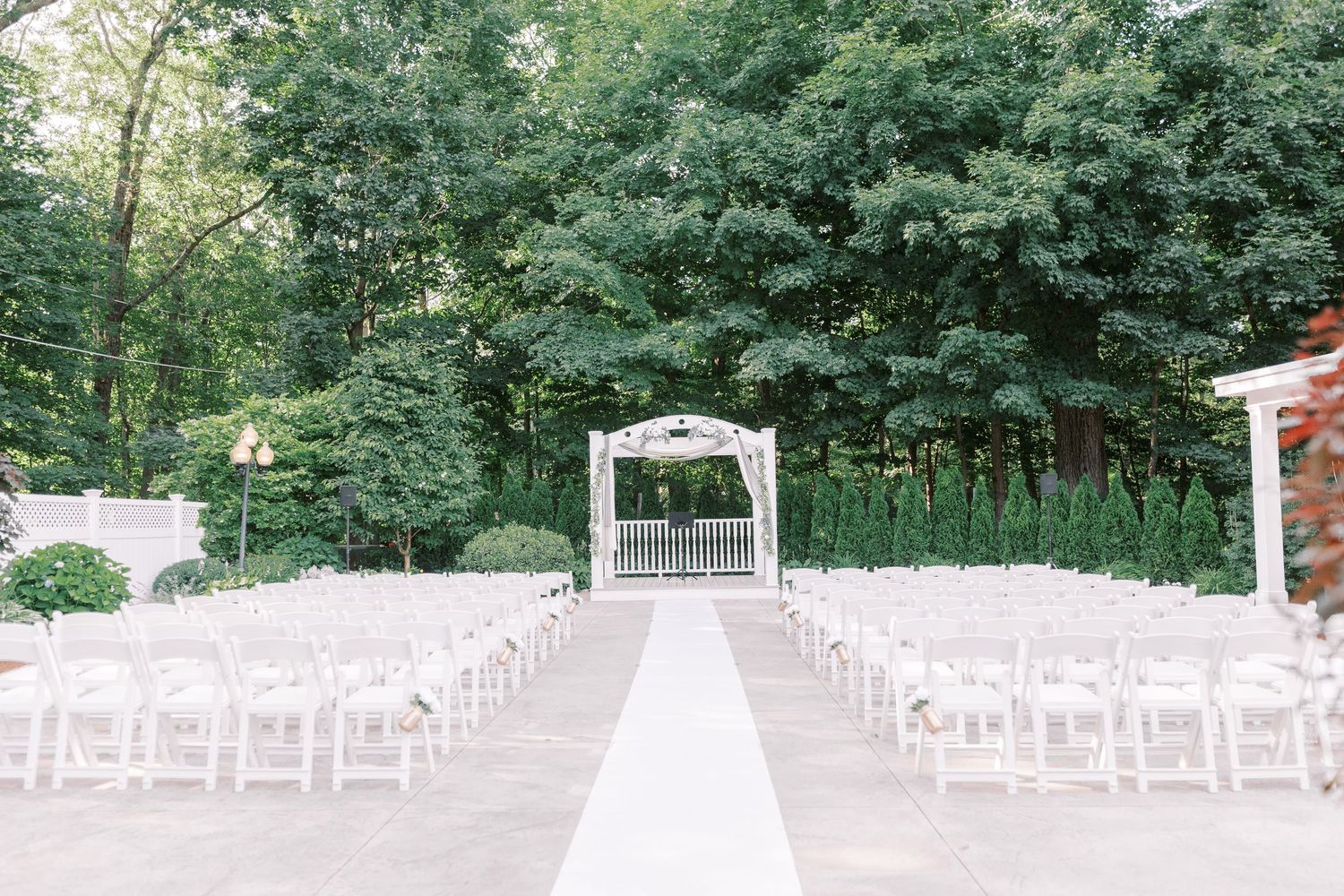 Elegant outdoor wedding ceremony setup with white chairs and a gazebo altar surrounded by lush green trees.