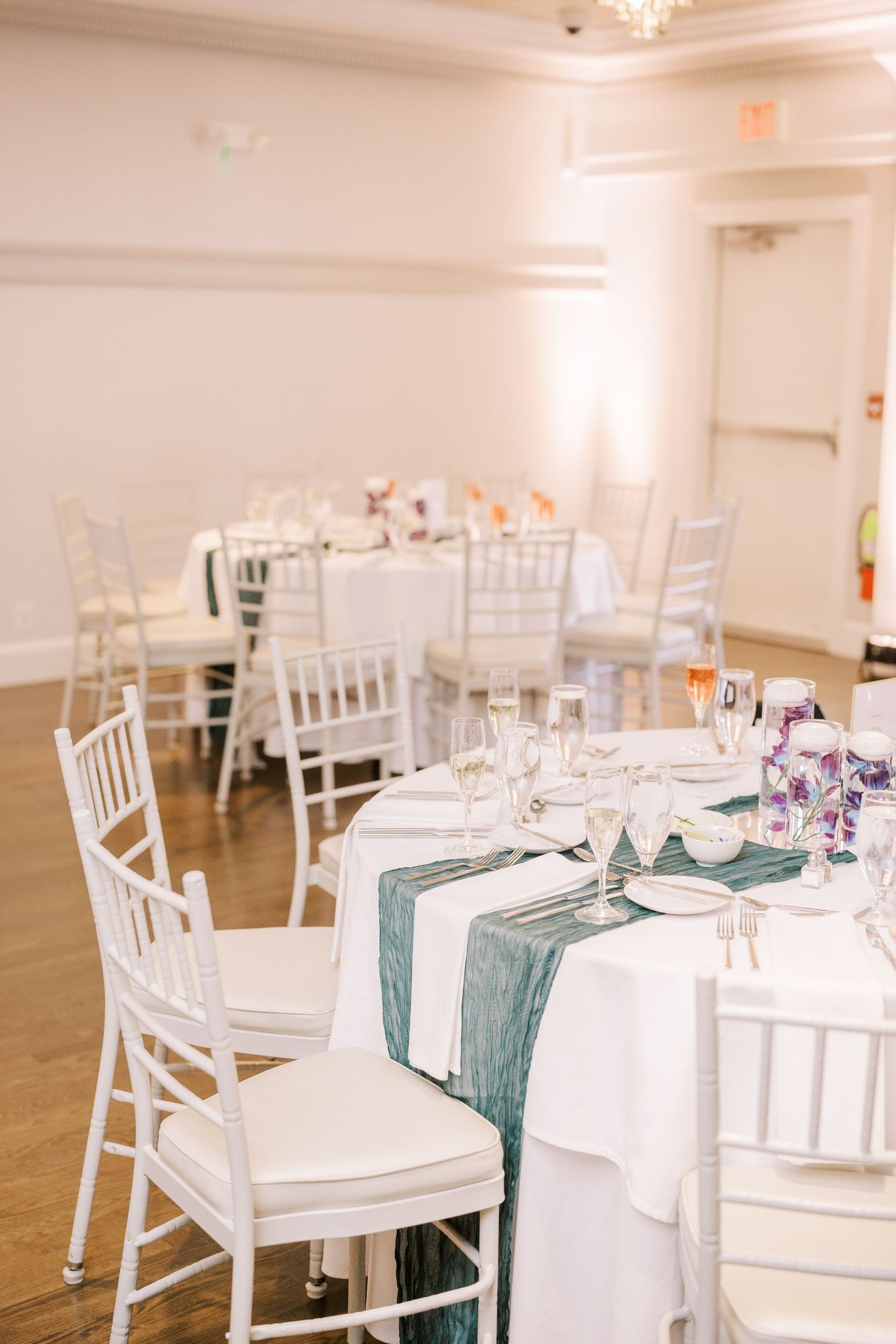 Elegant wedding reception tables with white chiavari chairs and teal table runners in a bright banquet room.