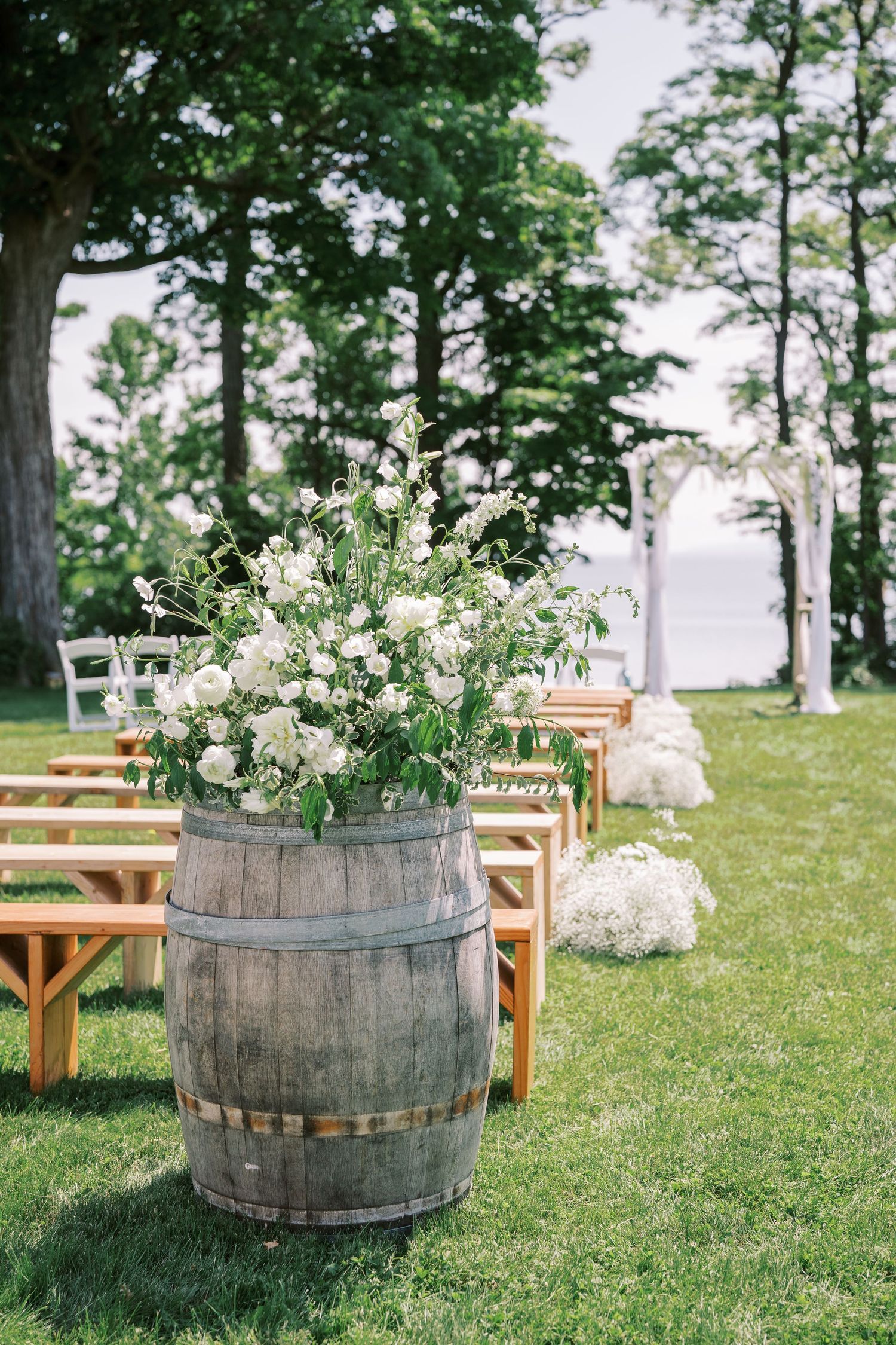 Rustic wooden barrel with white floral arrangement decorates outdoor wedding ceremony aisle setup by a lakeside.