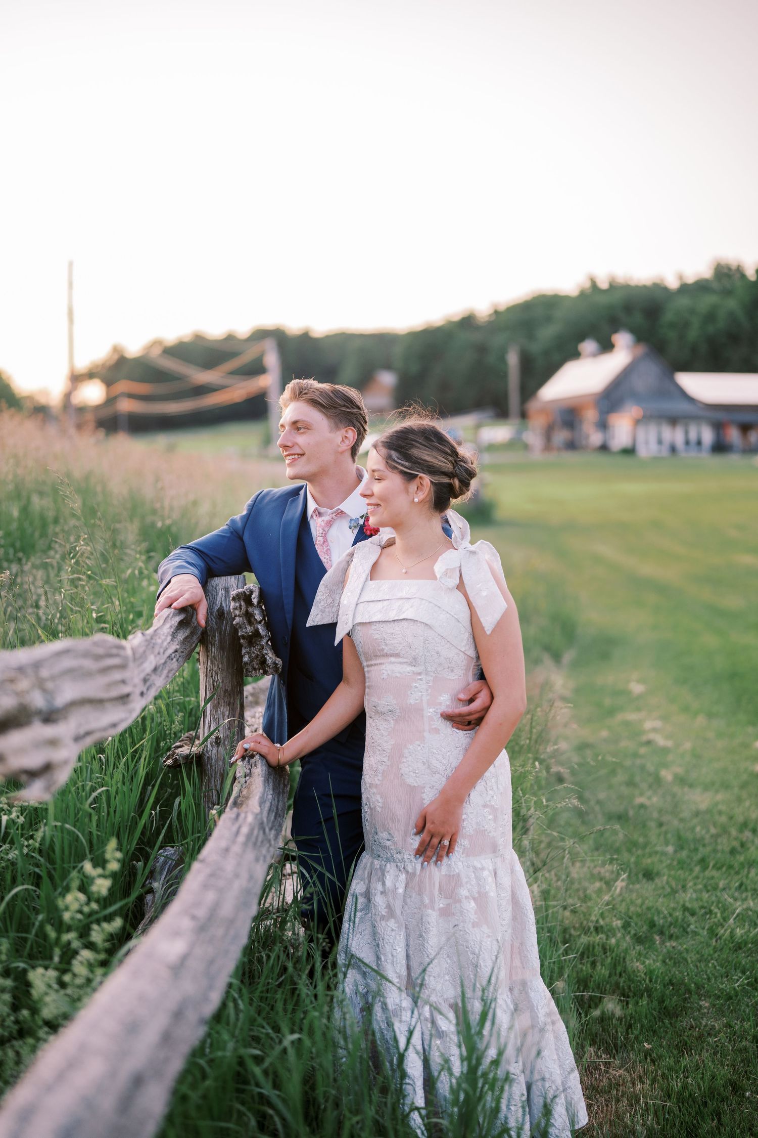 A couple in wedding attire stands together by a rustic wooden fence at sunset with a barn in the background.