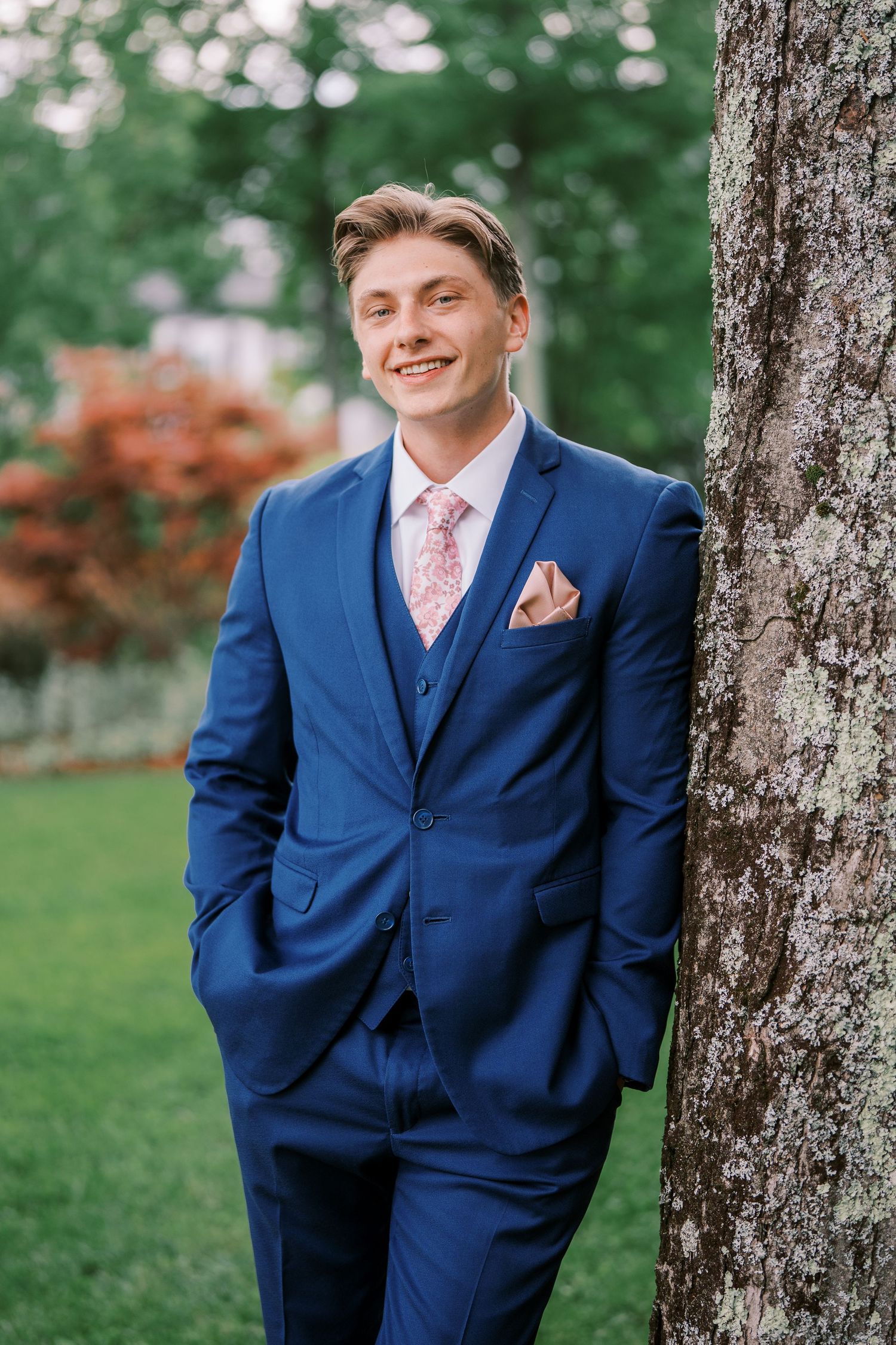 A person in a stylish blue three-piece suit with pink tie poses against trees in an outdoor portrait setting.