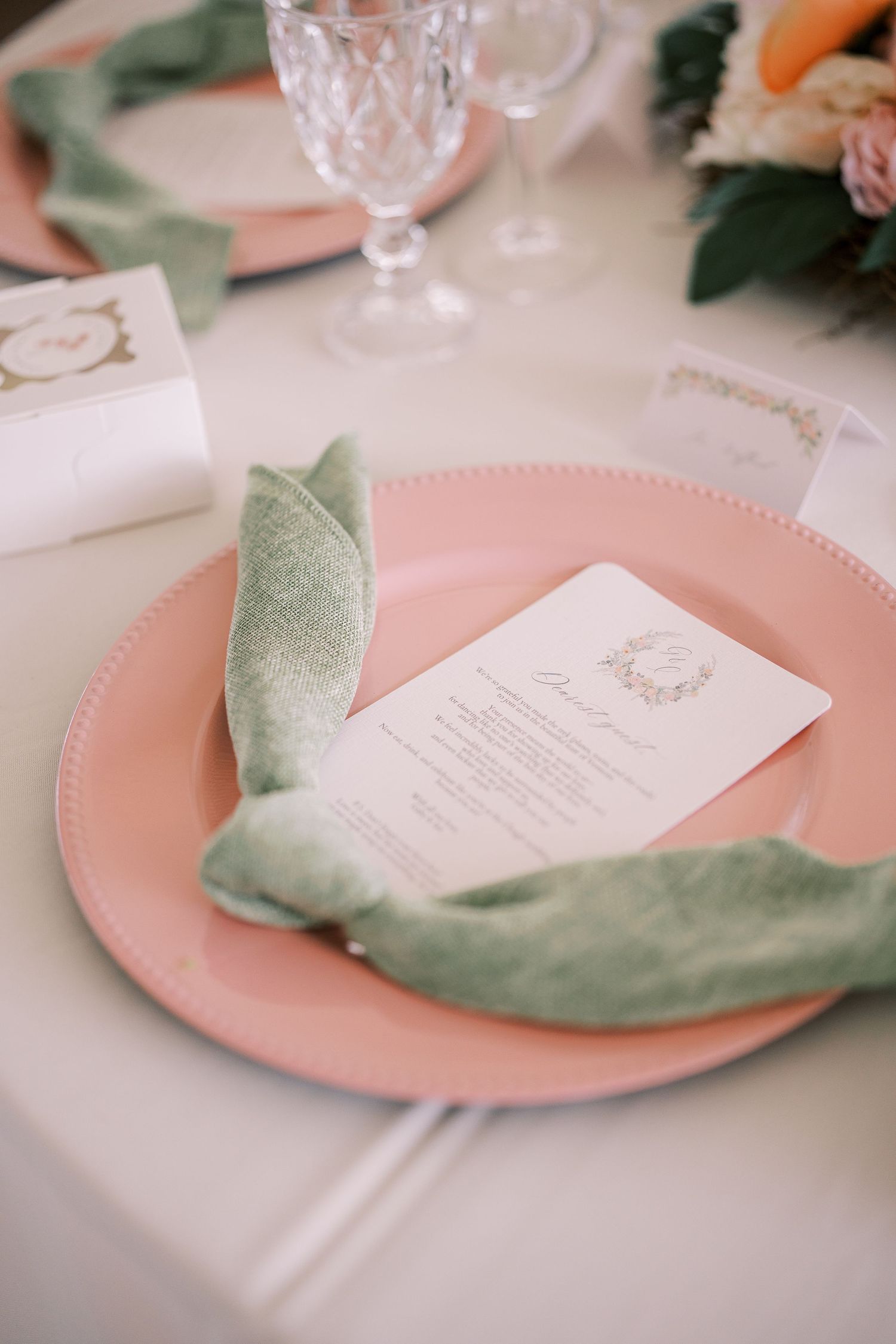 A wedding menu card rests elegantly on a blush pink charger plate with a soft sage green linen napkin at a reception table.