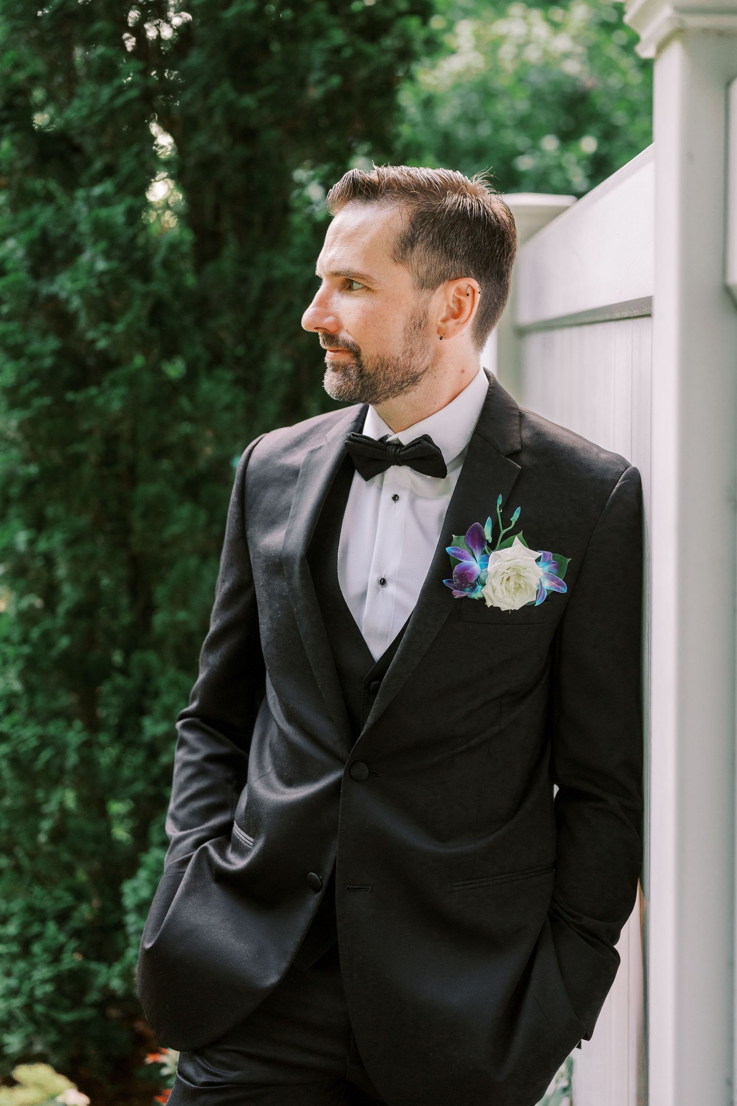 A groom in a black tuxedo and bow tie with a boutonniere looks to the side while standing by a white pillar outdoors.