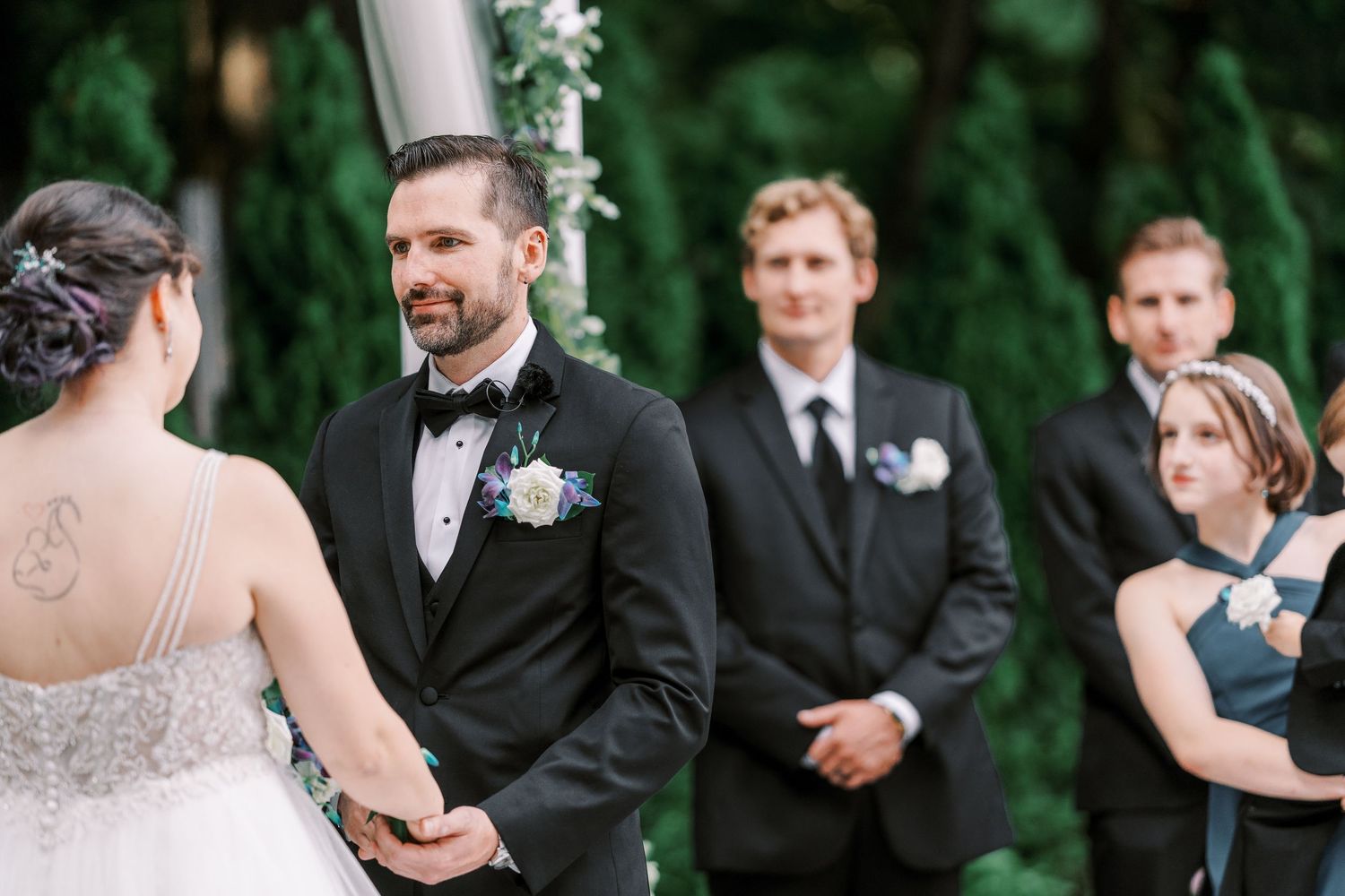 Wedding party stands outdoors on stone steps surrounded by lush green foliage during a summer celebration.