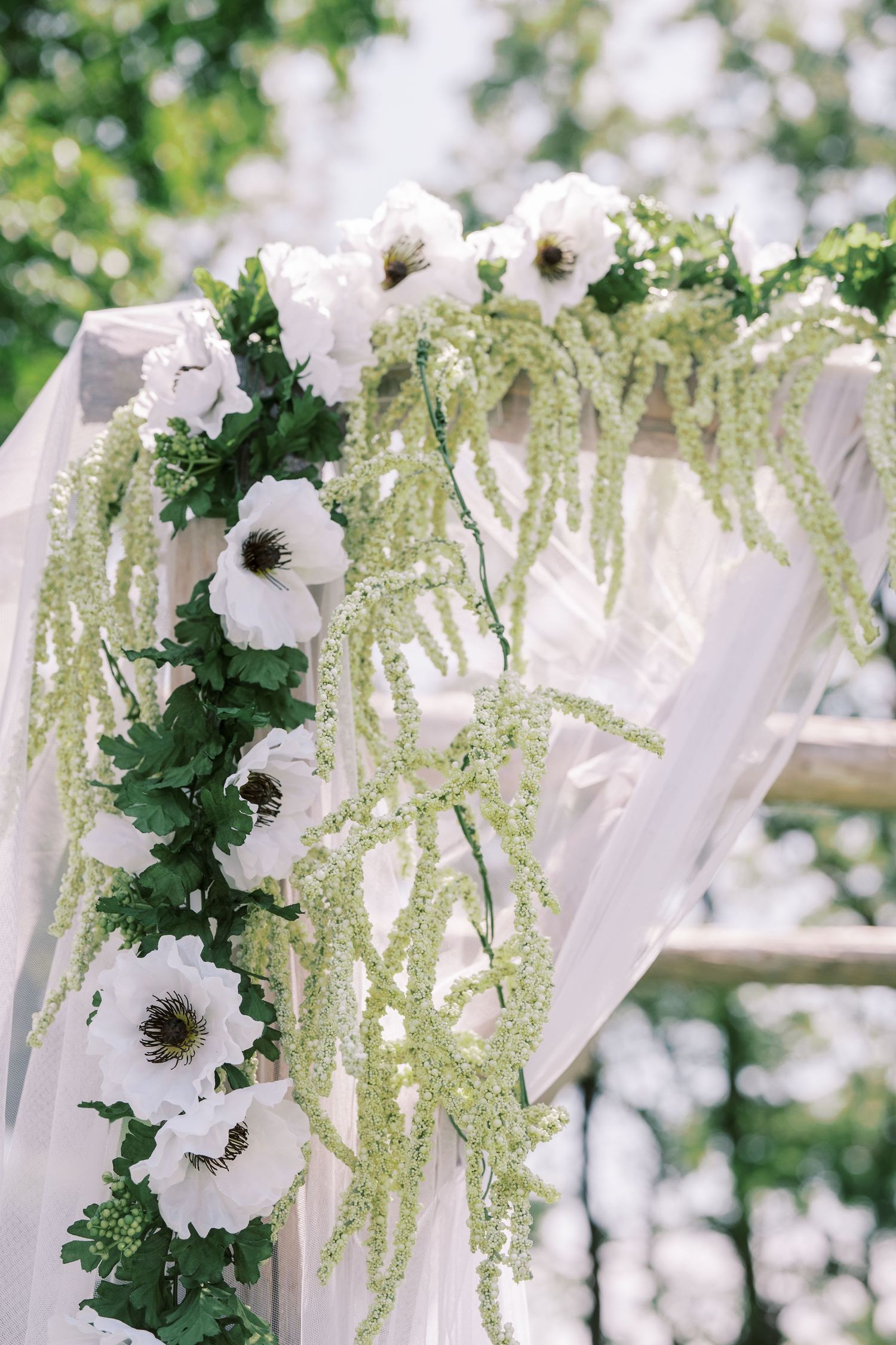 White floral wedding arch decoration with anemone flowers and cascading greenery against soft tulle draping outdoors.