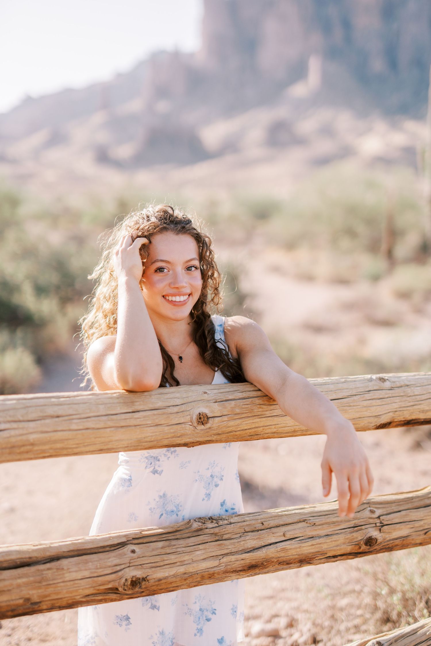Smiling woman in white floral dress leans against wooden fence railing in desert landscape at sunset.