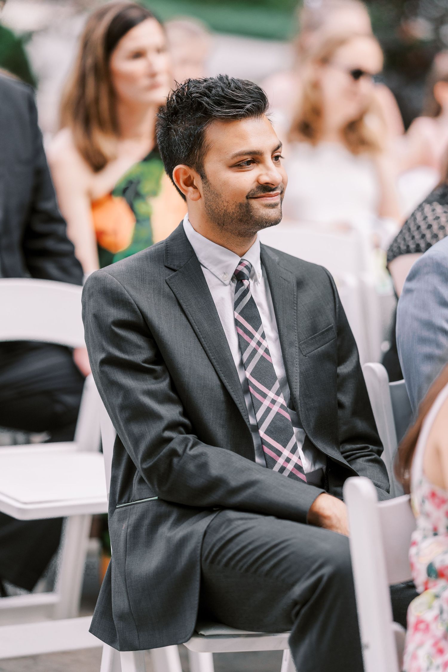 Wedding guest in dark suit and plaid tie sits attentively on white folding chair during outdoor ceremony.