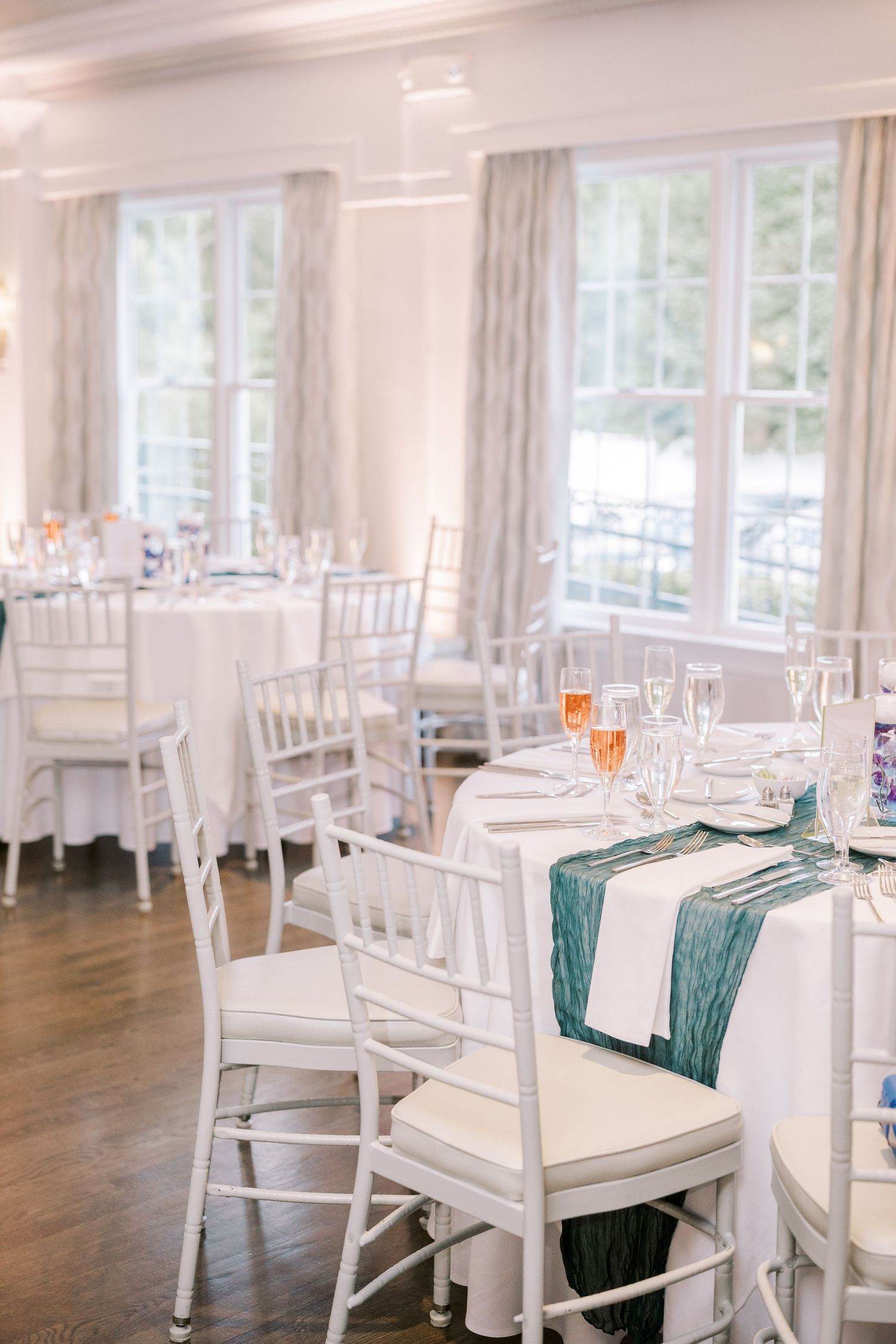 Elegant wedding reception venue with white chiavari chairs, round tables, and teal table runners in a bright room with windows.