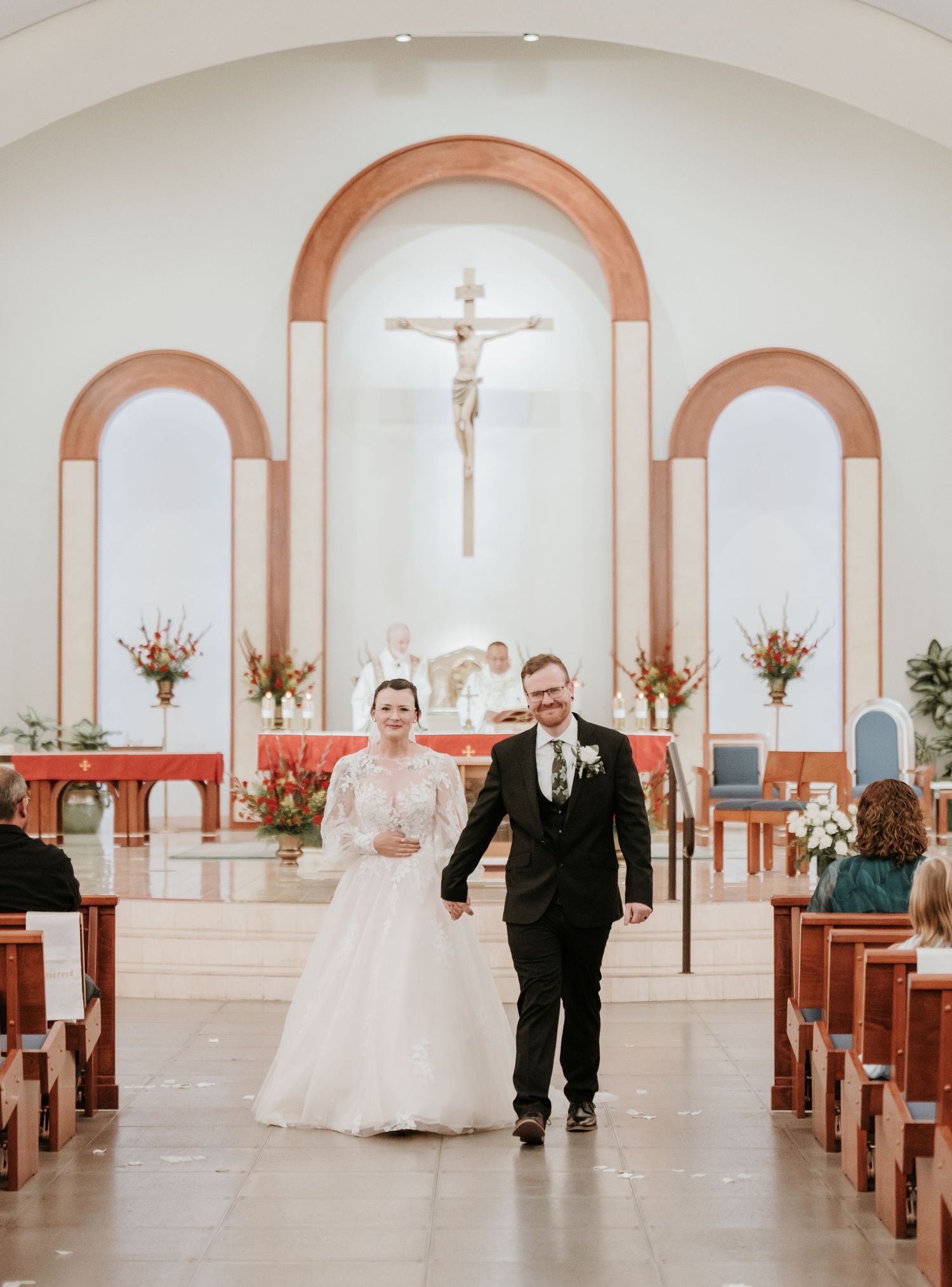 A couple walks down the aisle of a Catholic church with arched alcoves, crucifix, and floral arrangements at the altar.