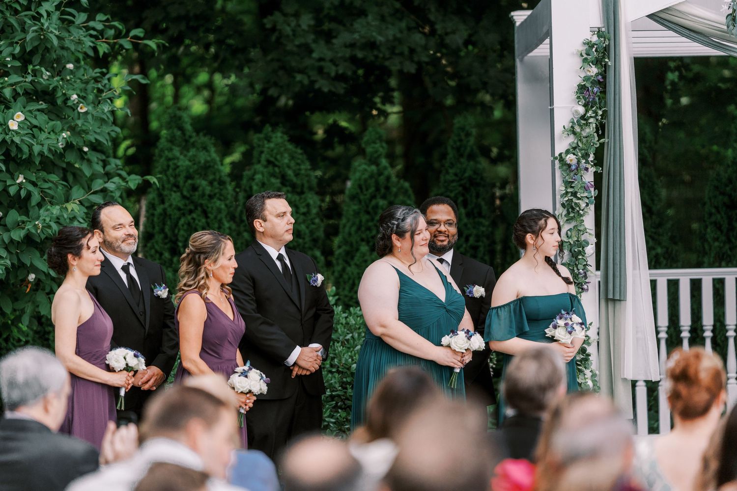 Wedding party gathers under a white pergola during an outdoor ceremony surrounded by lush green trees.