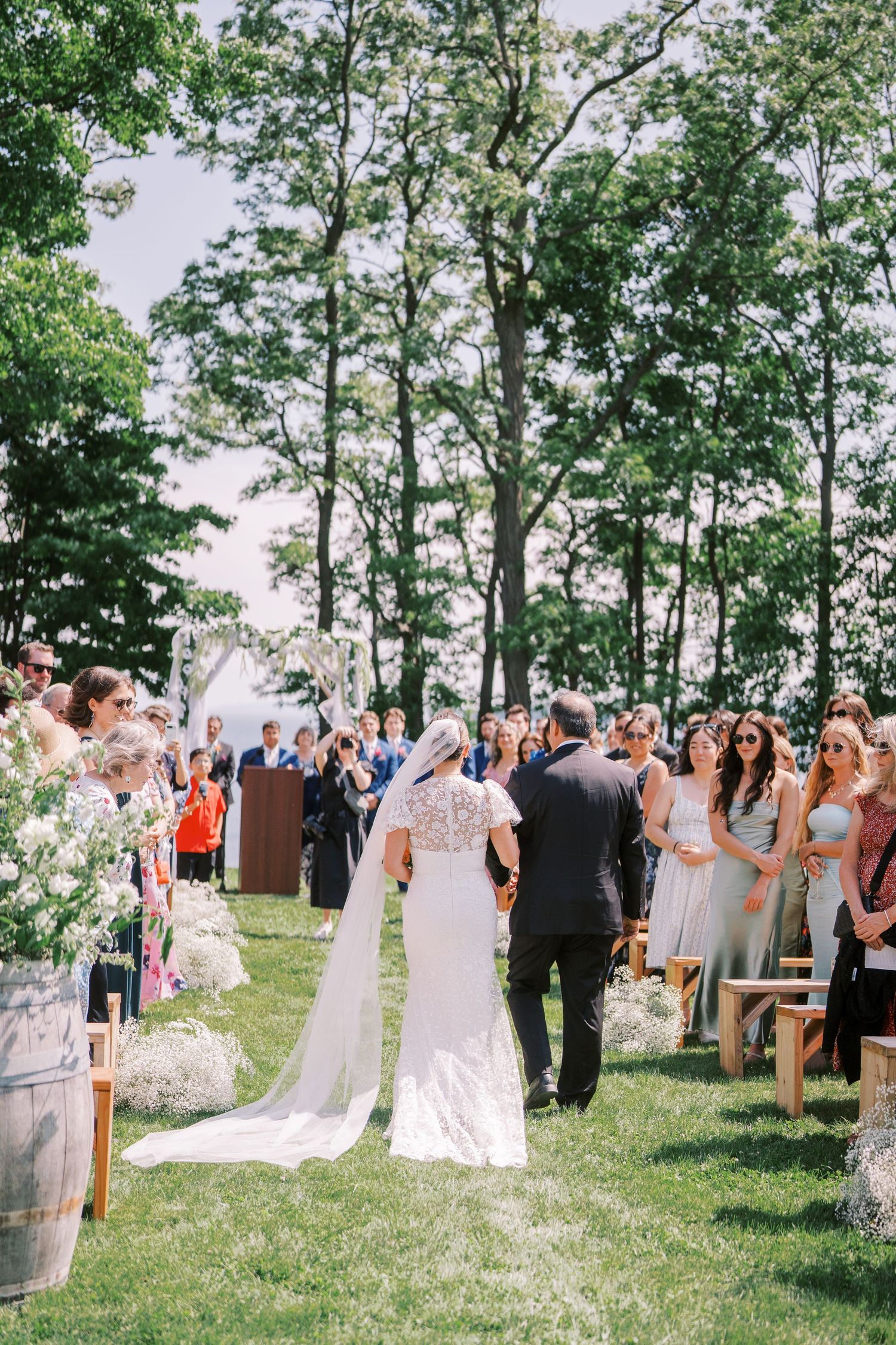 Wedding guests gather in an outdoor ceremony space surrounded by tall trees on a sunny spring afternoon.