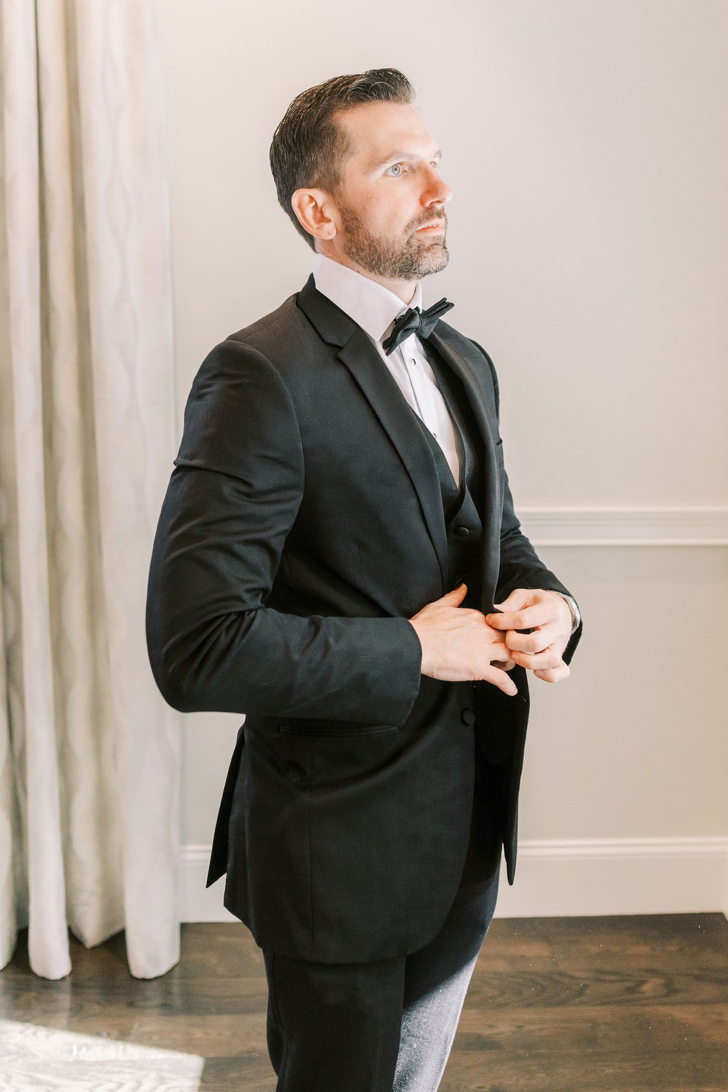 Side profile of a formal black tuxedo and bow tie being worn against a light wall with floor-length curtains.