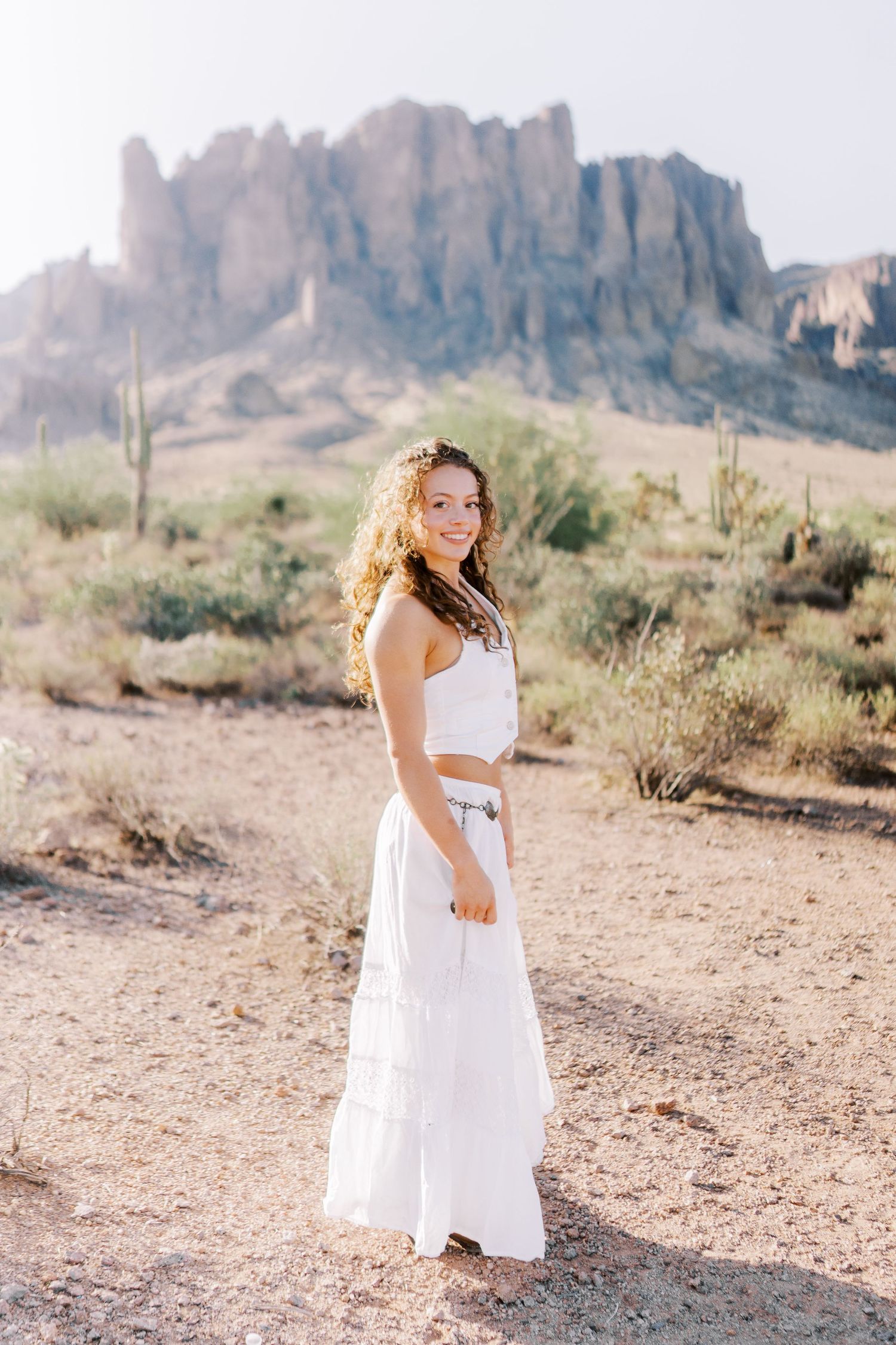A white maxi dress worn in a desert landscape with rugged mountains and saguaro cacti in the background.