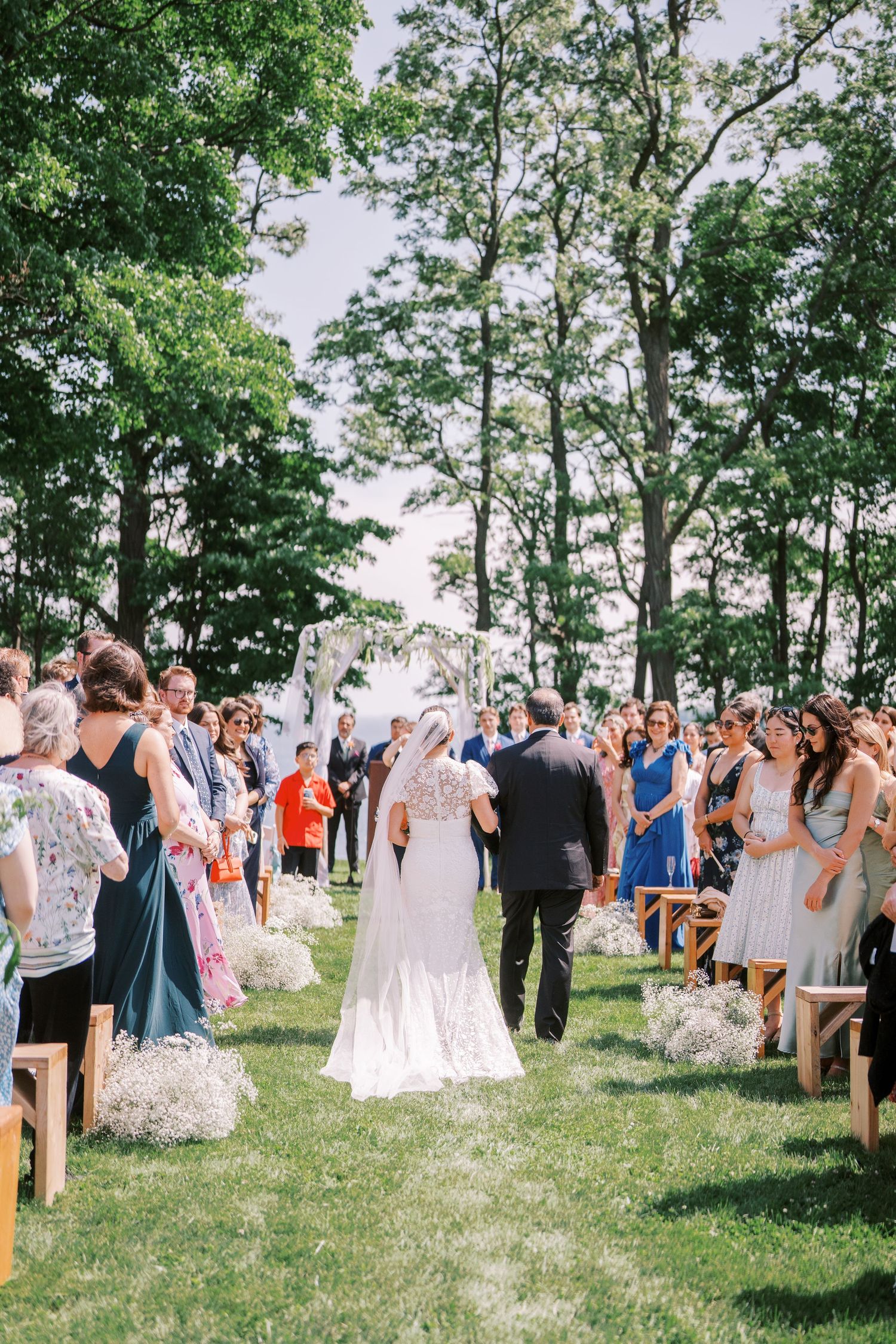 An outdoor wedding ceremony takes place on a grassy lawn surrounded by tall trees as guests gather on wooden chairs.