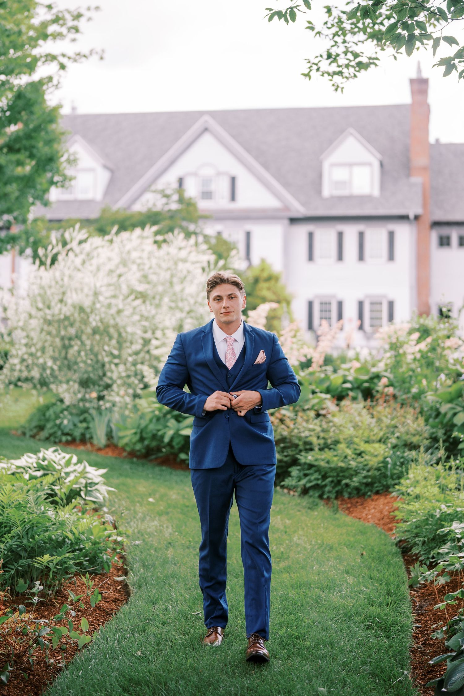 A person in a navy blue suit and tie stands on a garden path with a white colonial house in the background.