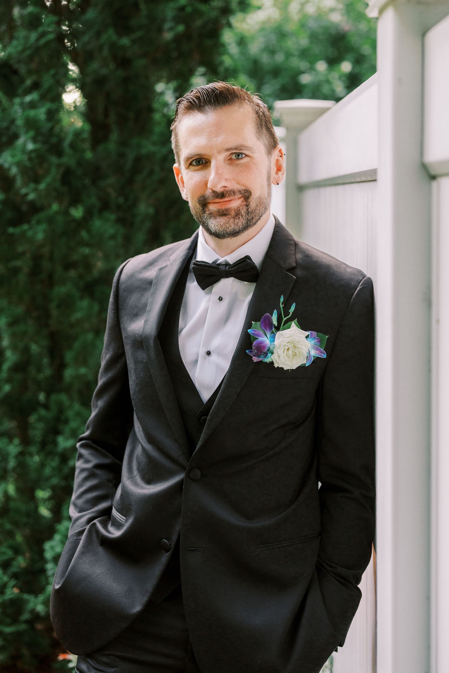 Groom in black tuxedo with bow tie and floral boutonniere leans against white pillar outdoors.