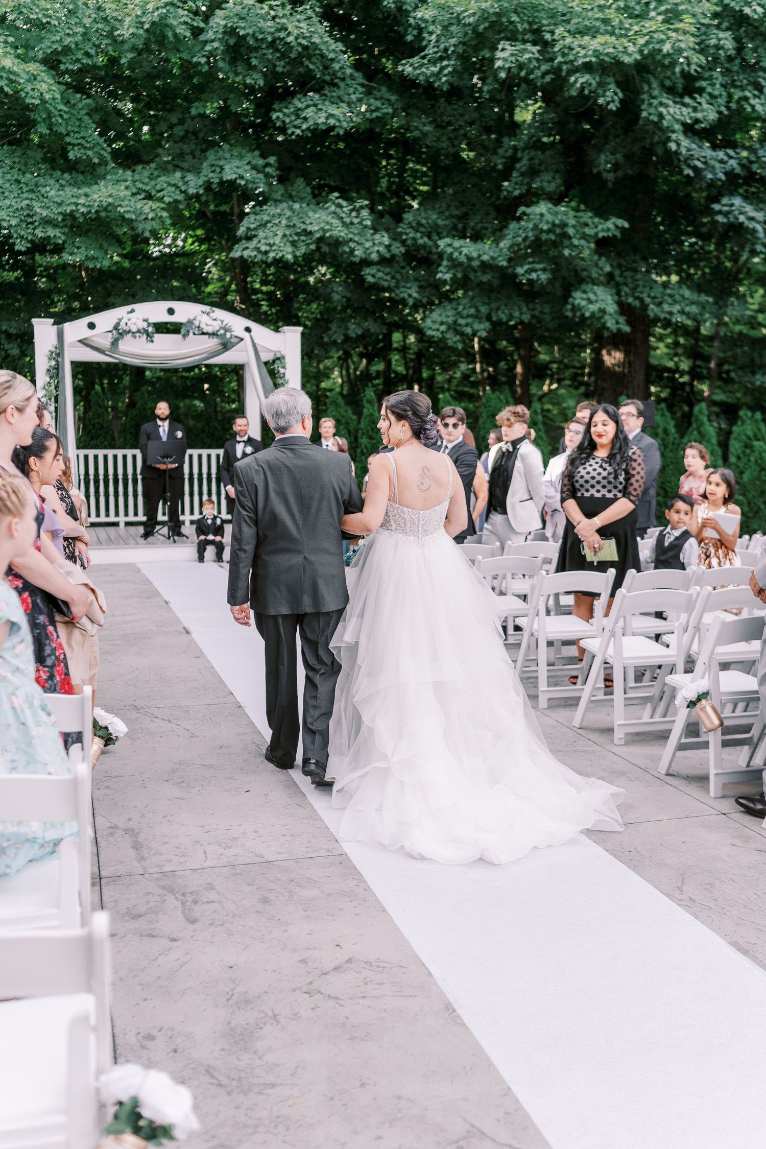 Outdoor wedding ceremony at a garden venue with white chairs and floral arch along a white aisle runner.
