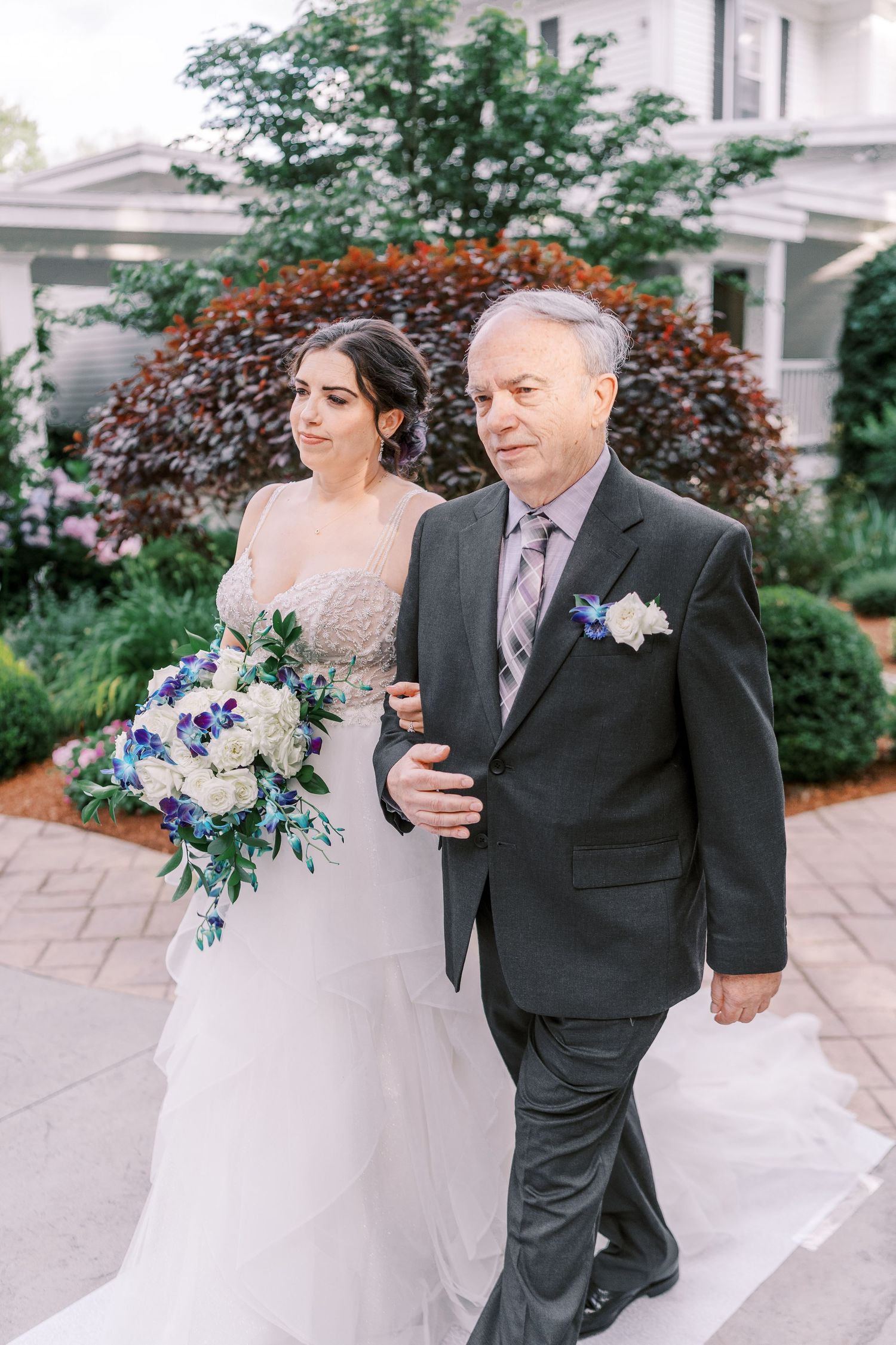 An elegant wedding procession on a paved path surrounded by lush gardens and landscaping with blue floral bouquet.