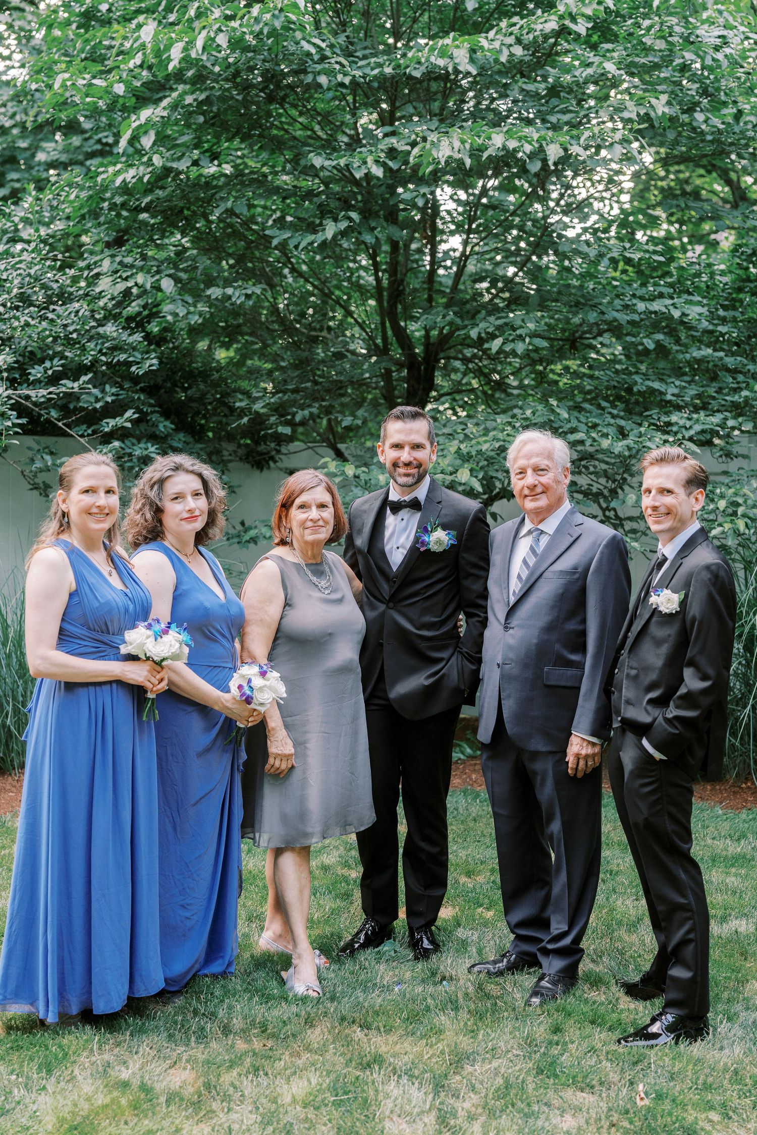 Group in formal attire poses on a lawn with lush green trees forming a natural backdrop at an outdoor event.
