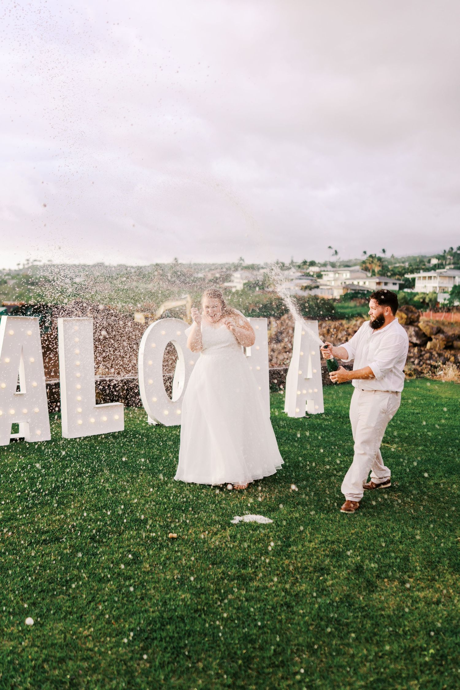 Wedding couple celebrates with champagne spray in front of large ALOHA letters on a grassy lawn overlooking coastal hills.