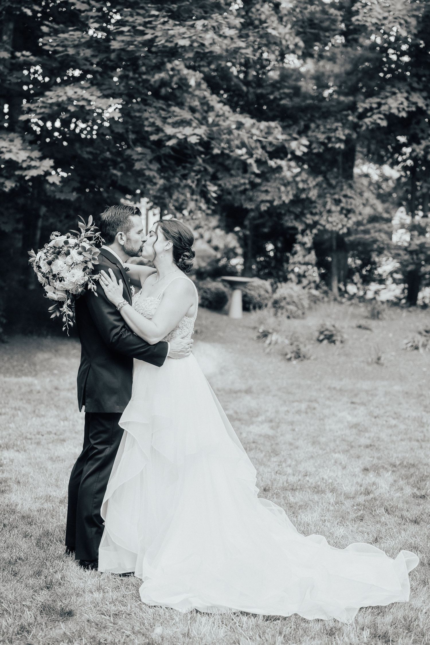A romantic black and white wedding photo showing a couple embracing on a grassy lawn surrounded by trees.