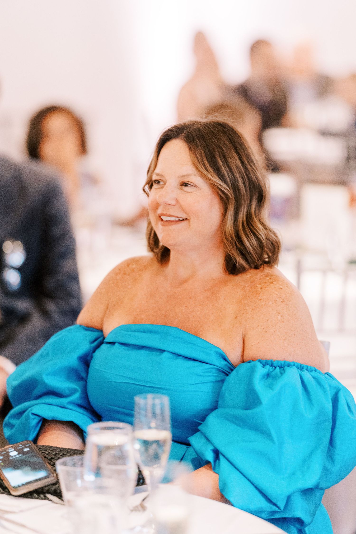 Guest in vibrant turquoise off-shoulder dress smiles while seated at formal event with table settings.
