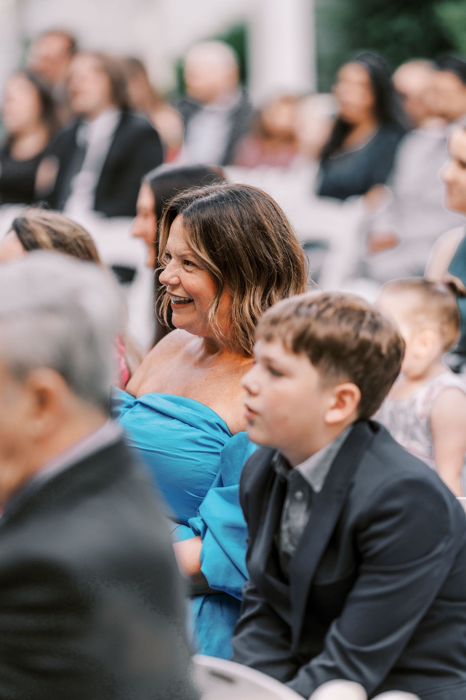 Wedding guests seated outdoors share joyful reactions during ceremony, with someone in turquoise dress and formal attire.