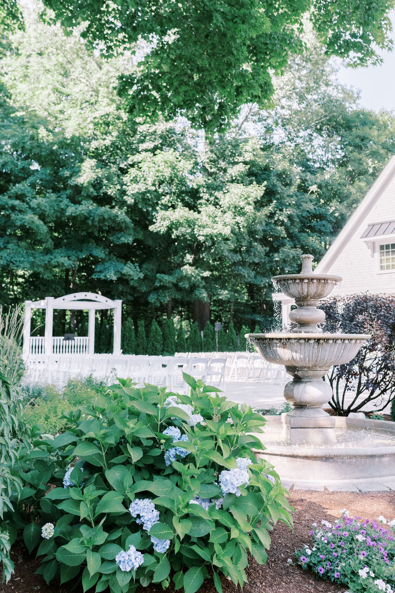 A serene garden pathway lined with blooming hydrangeas and decorative urns leads to a white gazebo in dappled sunlight.