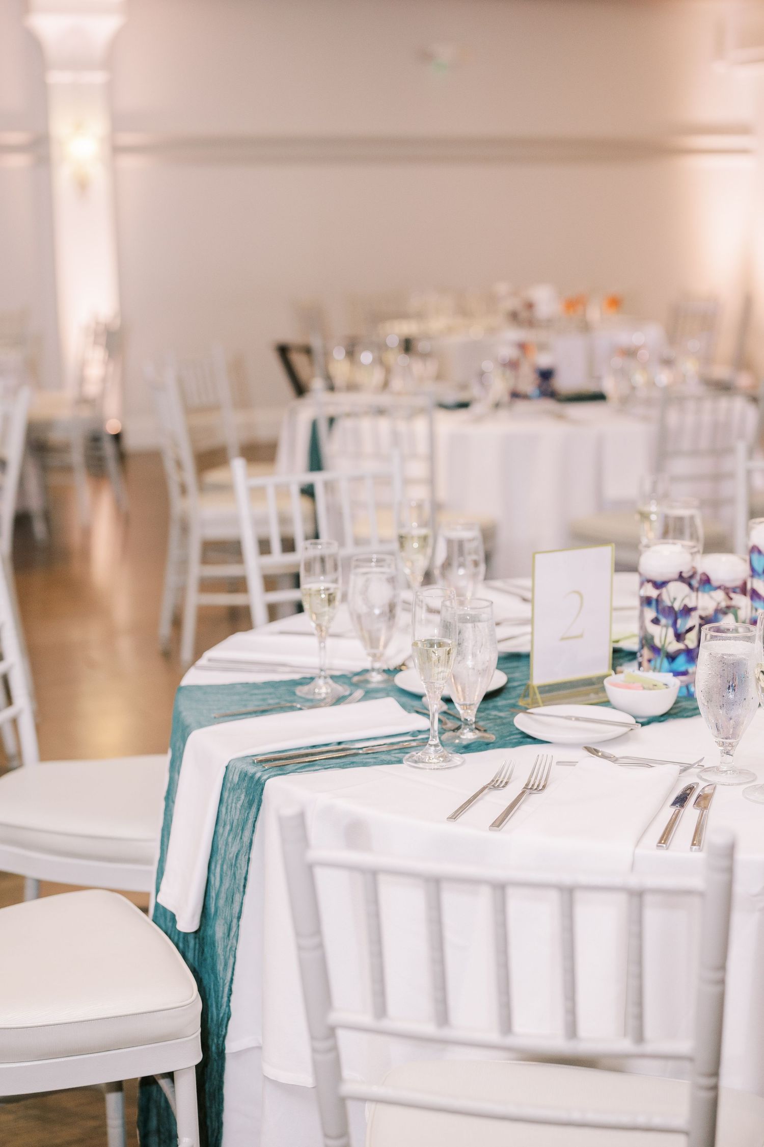 Elegant table setting with white linens, teal runners and white chiavari chairs in a wedding reception venue.