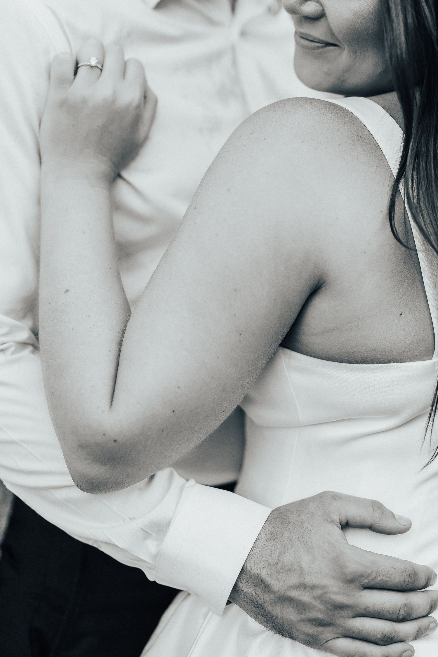Black and white close-up of two people embracing in formal white attire, with hands tenderly placed on each other.