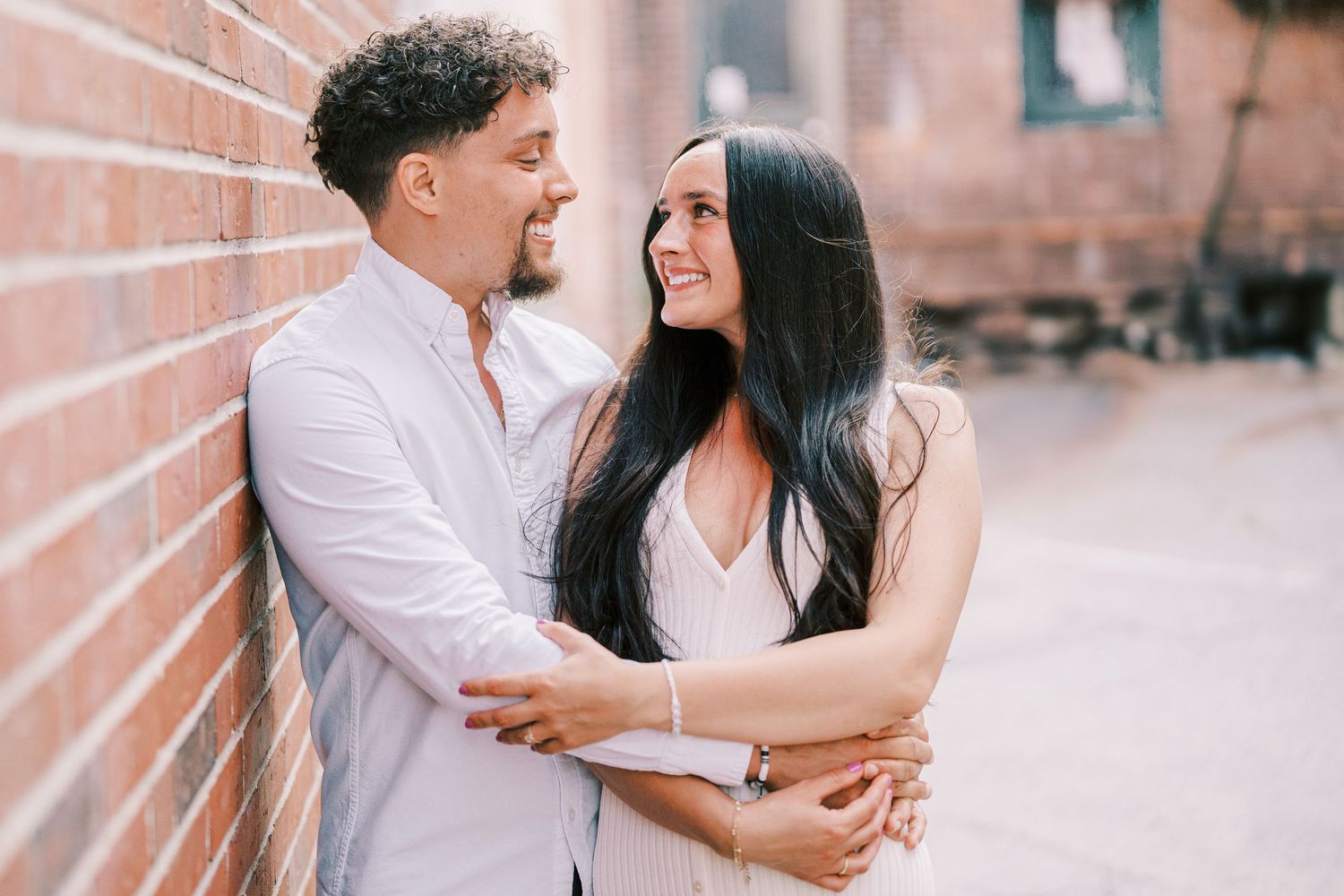 A happy couple embraces and laughs together while leaning against a rustic brick wall outdoors.