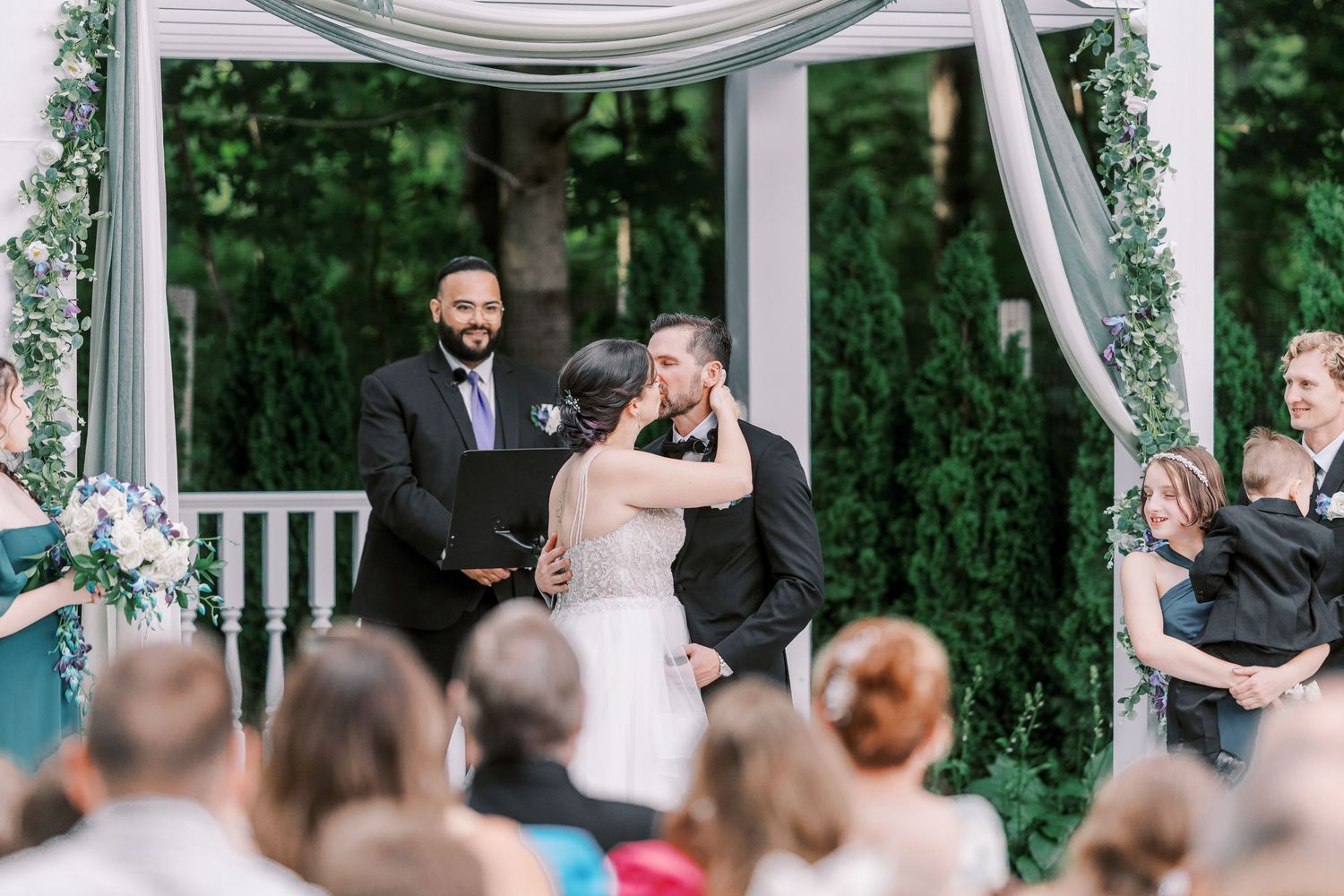 Wedding party stands under an elegant white draped gazebo during an outdoor garden ceremony.